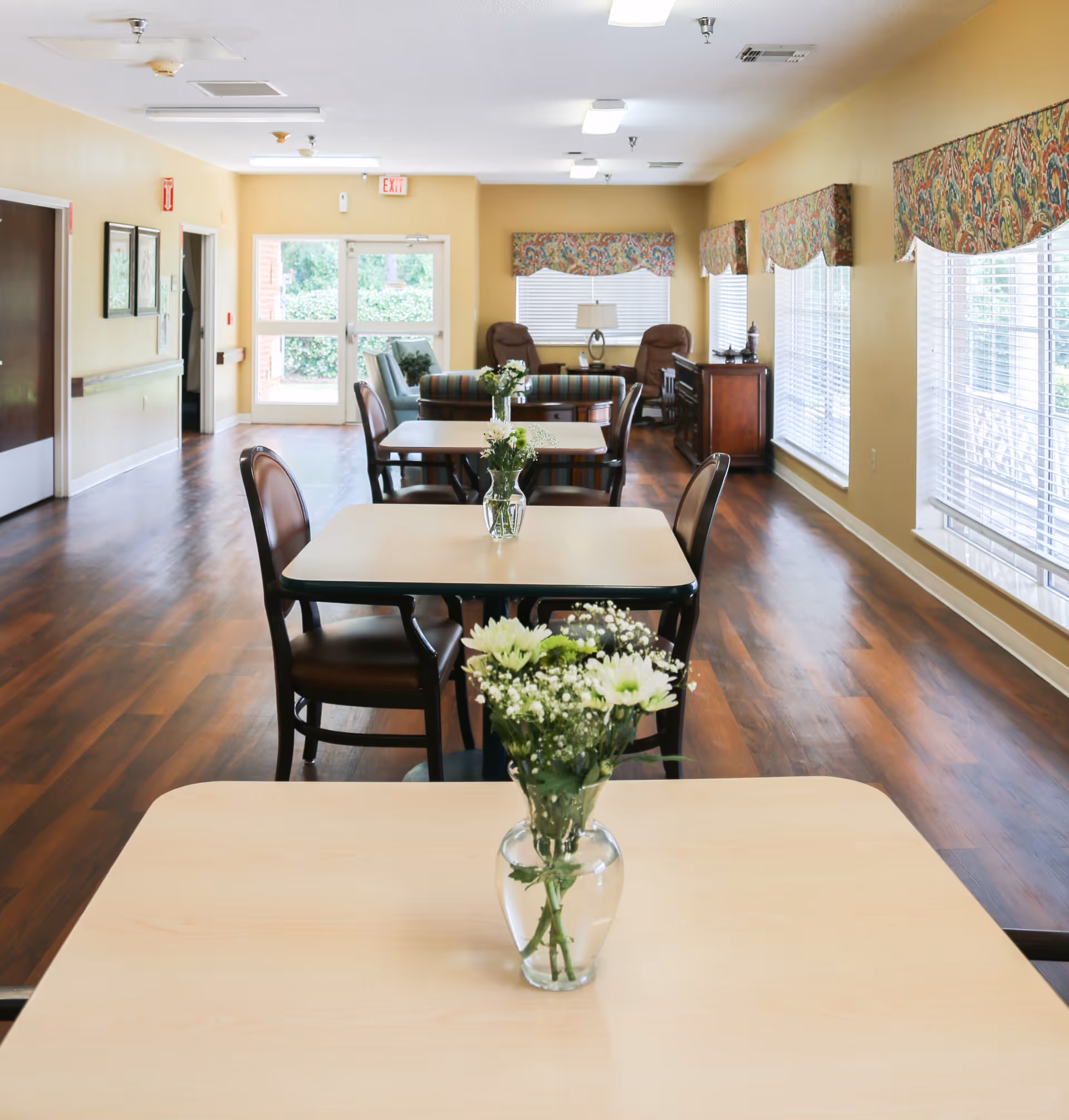 Bright senior dining room with rows of tables and chairs, vases of flowers, wooden floors and a seating area by the windows.