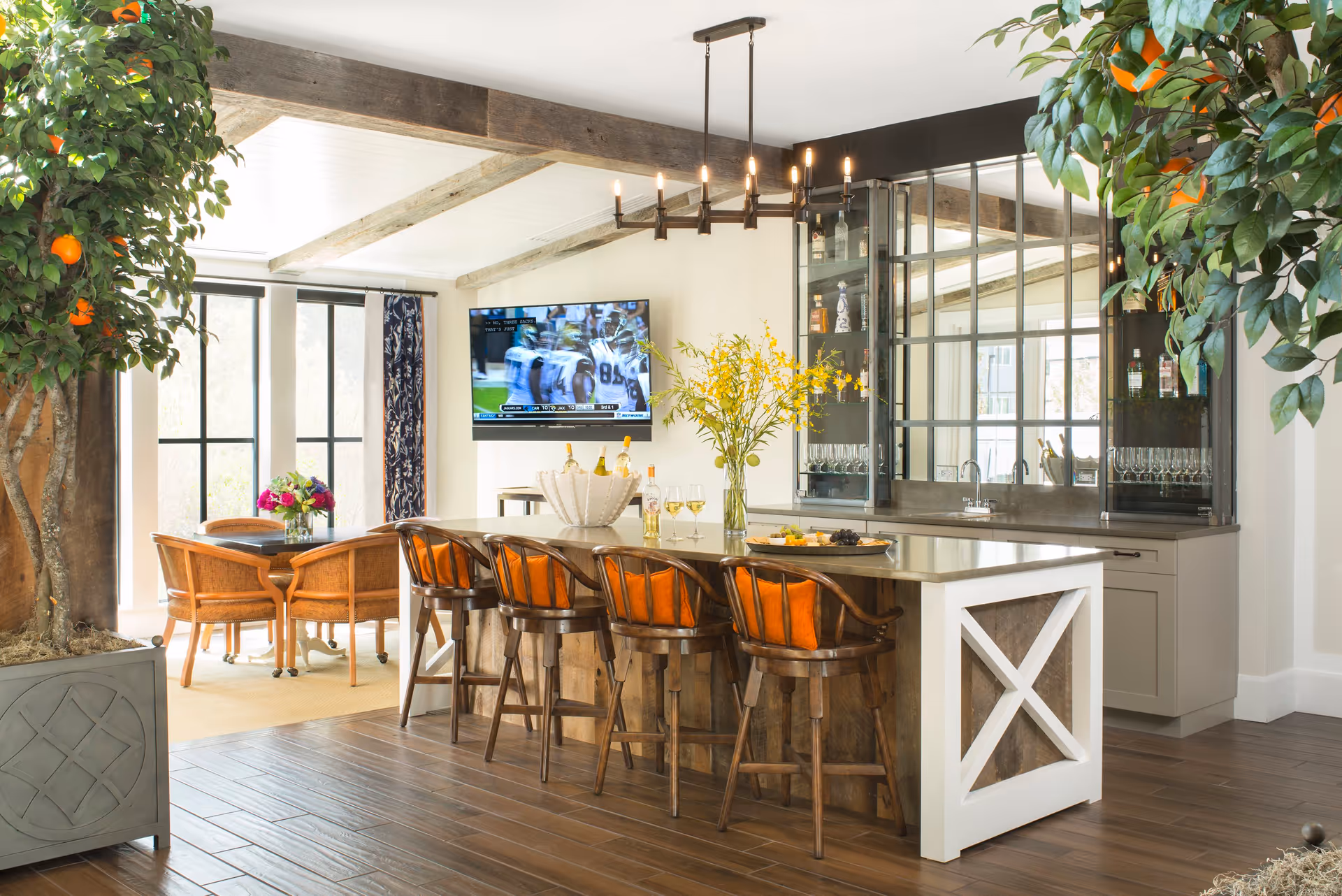 A bright and spacious interior area featuring a kitchen island with five wooden bar stools with orange cushions. The island has a vase with yellow flowers, wine glasses, and a bottle. Behind the island is a countertop with a sink and glass cabinets. A flat-screen TV is mounted on the wall showing a sports game. To the left, there is a small dining area with a round table and four wooden chairs, with a flower arrangement on the table. Large windows let in natural light, and there are two large potted orange trees framing the scene. The ceiling has exposed wooden beams and a modern chandelier above the island.