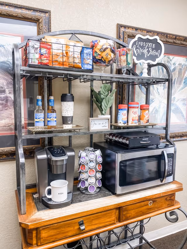 A coffee and snack station with a metal rack on a wooden table. The top shelf holds baskets with various snacks including chips and crackers. The middle shelf has coffee creamers, syrup bottles, a stack of disposable coffee cups, and a small potted plant with a sign that reads 'You can do it! coffee'. The bottom shelf features a Keurig coffee maker with a mug labeled 'sip.', a rotating stand with coffee pods, and a microwave oven. Behind the station are framed paintings and a small sign that says 'Employee Break Room'.