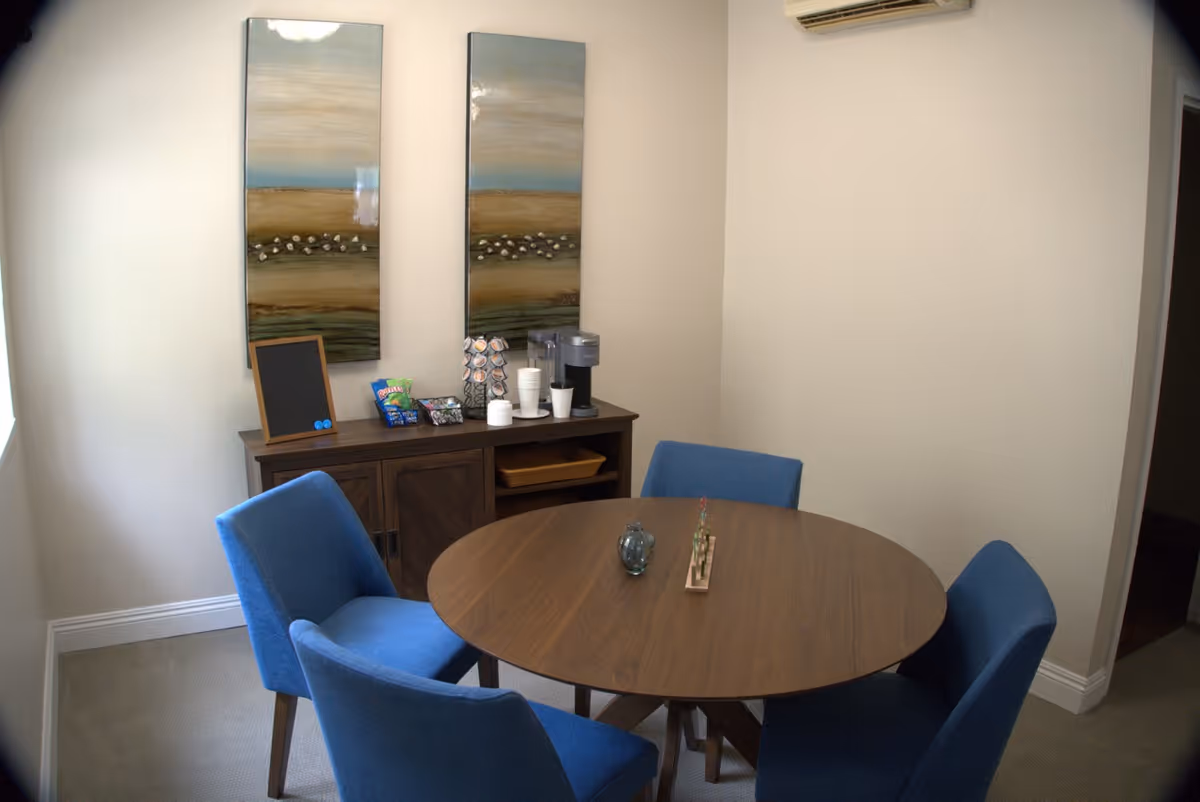 A small dining area with a round wooden table surrounded by four blue upholstered chairs. Against the wall is a wooden sideboard with a coffee maker, disposable cups, snacks, and a small chalkboard. Two vertical landscape paintings hang above the sideboard. The room has beige walls and carpeted floor.