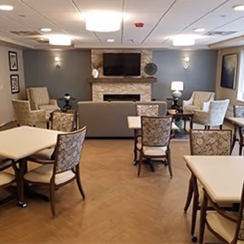 A cozy common area with multiple tables and chairs arranged for seating. The room features a stone fireplace with a mounted flat-screen TV above it, flanked by decorative items. Comfortable armchairs and a sofa are positioned around the fireplace, and the walls are painted gray with framed artwork and wall sconces providing soft lighting.