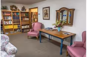 A cozy common room with bookshelves, upholstered chairs, a wooden console table with a flower arrangement, and a wall mirror.