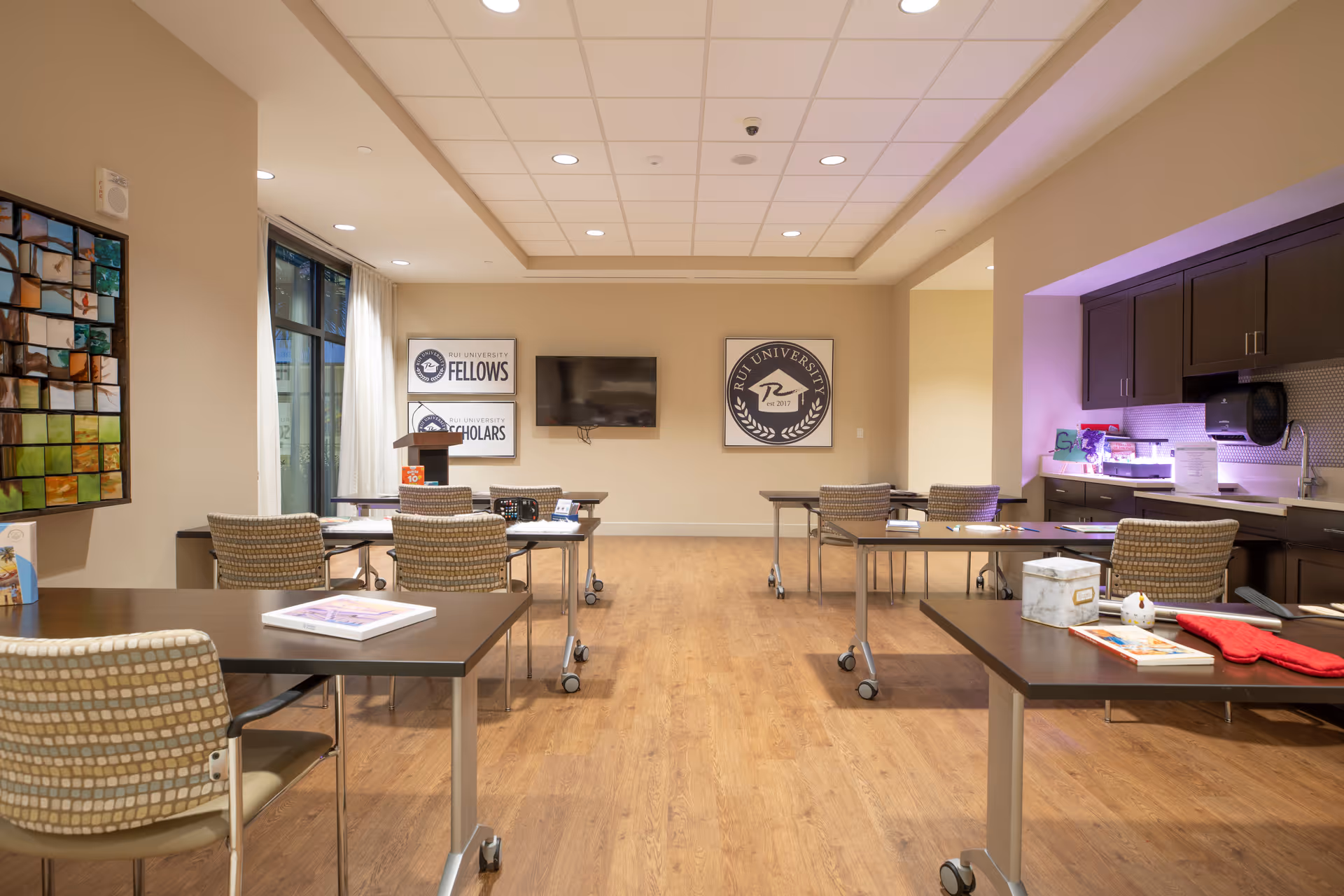 Well-lit multi-purpose activity room with tables and chairs, a wall-mounted TV, artwork, and a kitchenette area.