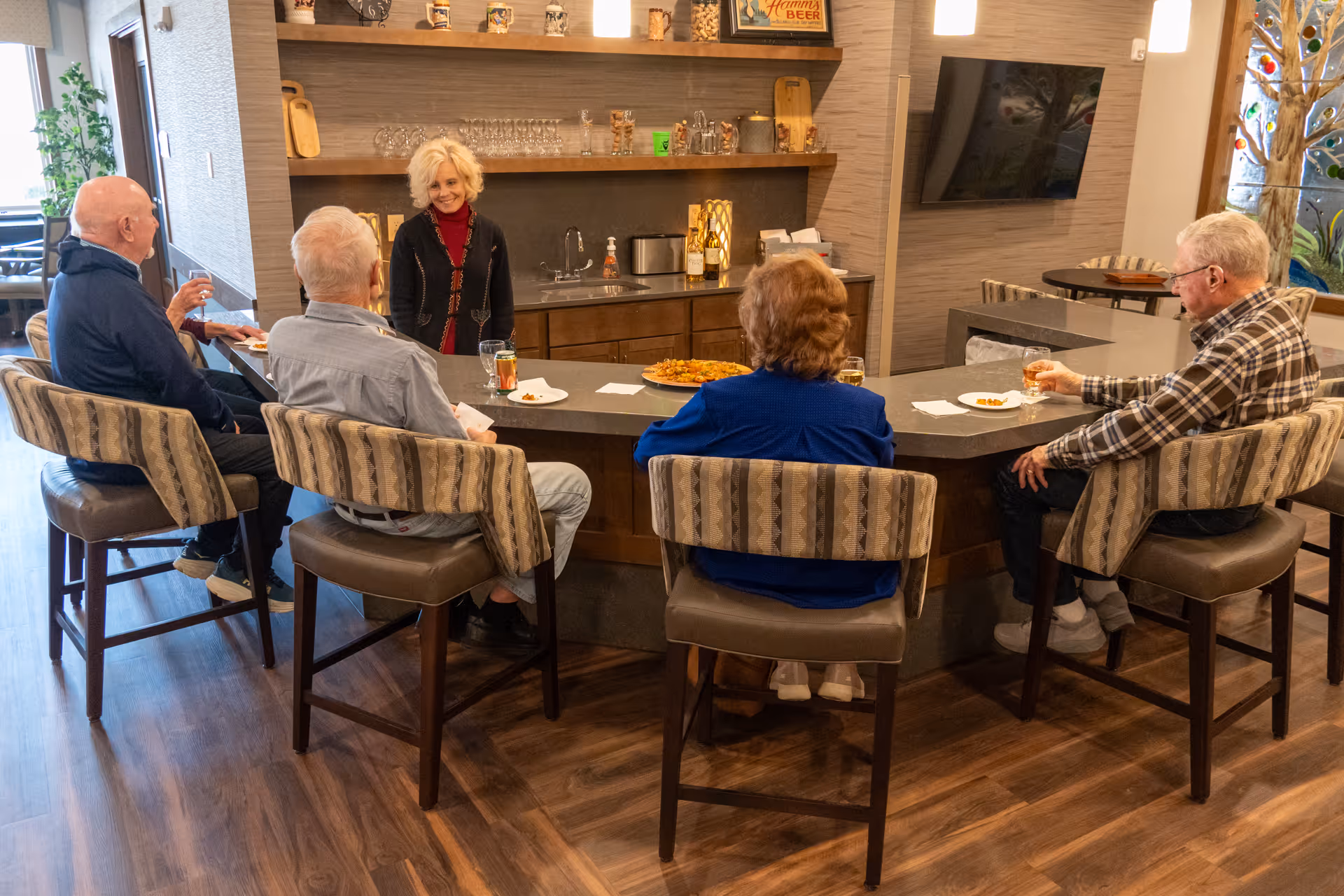 A group of four elderly people sitting on bar stools around a kitchen island counter, enjoying drinks and snacks, while a woman stands behind the counter smiling. The room has wooden flooring, shelves with glassware and decorative items, and a wall-mounted TV.
