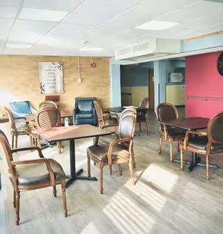 A bright room with several wooden tables and chairs arranged for seating. The room has light-colored wooden flooring, a brick accent wall with a framed decorative sign, and a mix of blue and brown chairs. Sunlight streams in through windows, casting shadows on the floor.
