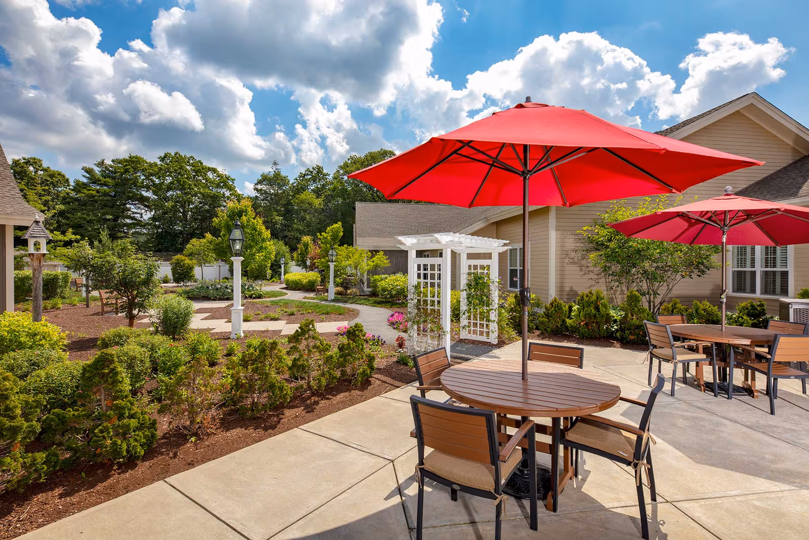 Outdoor patio and landscaped courtyard with round tables, red umbrellas, a pergola, and surrounding greenery by a building.
