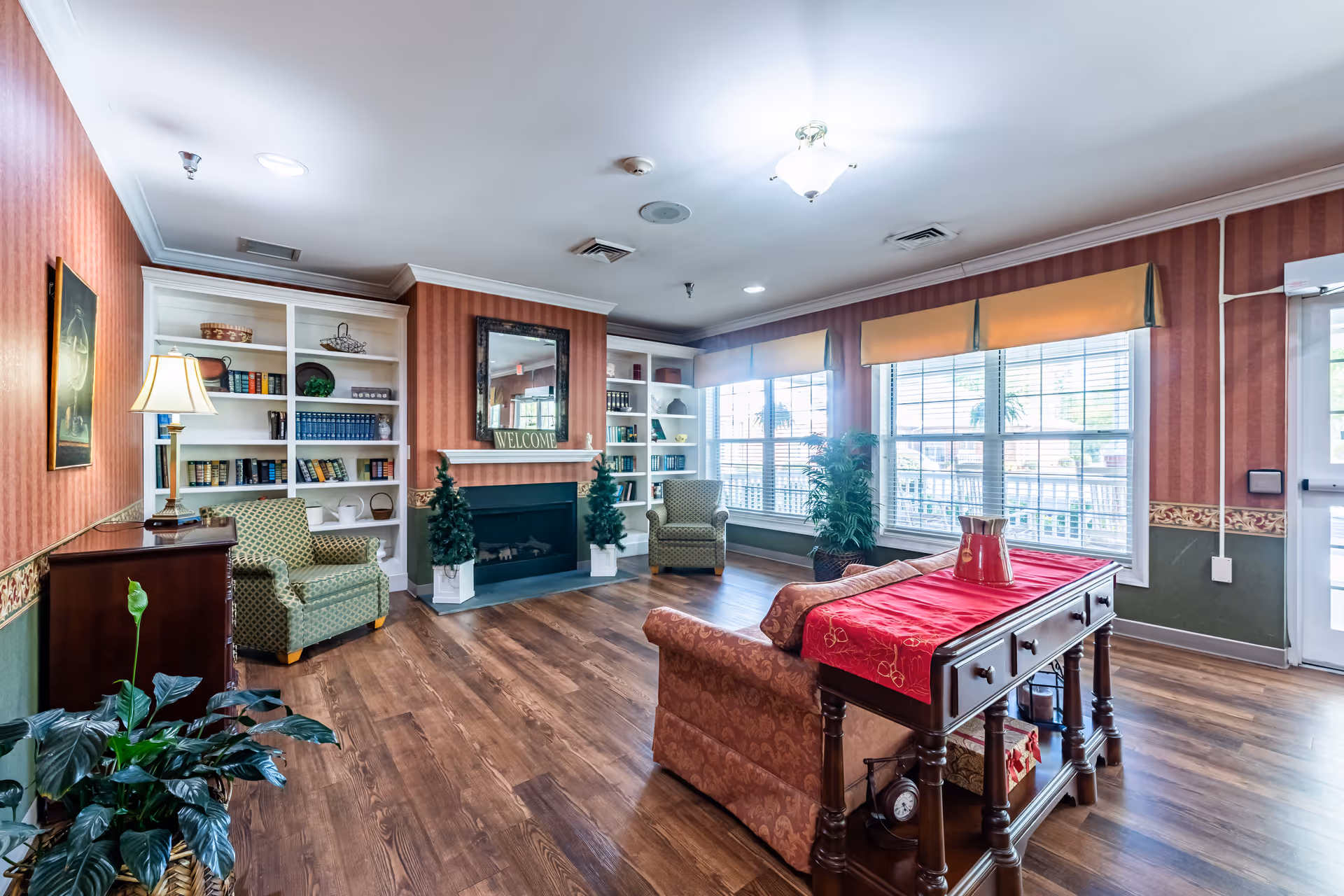 A cozy assisted living facility common area with wood flooring, patterned wallpaper, and large windows letting in natural light. The room features a fireplace with a 'WELCOME' sign above it, two green armchairs, a sofa with a red runner on the back, a wooden console table with decorative items, built-in white bookshelves filled with books and decor, and several potted plants.