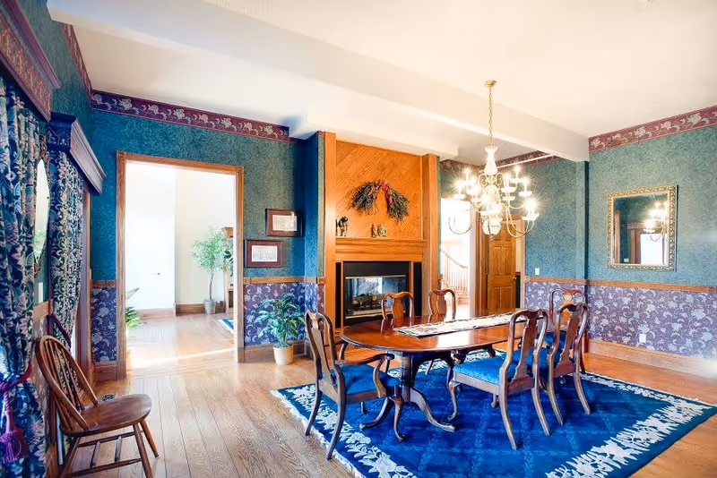 A traditional dining room with a wooden dining table and six chairs with blue cushions, a blue patterned rug underneath, a wooden fireplace with a decorative wreath above it, green and purple wallpaper with a floral border, a chandelier hanging from the ceiling, a mirror on the wall, and a doorway leading to a bright hallway with plants.