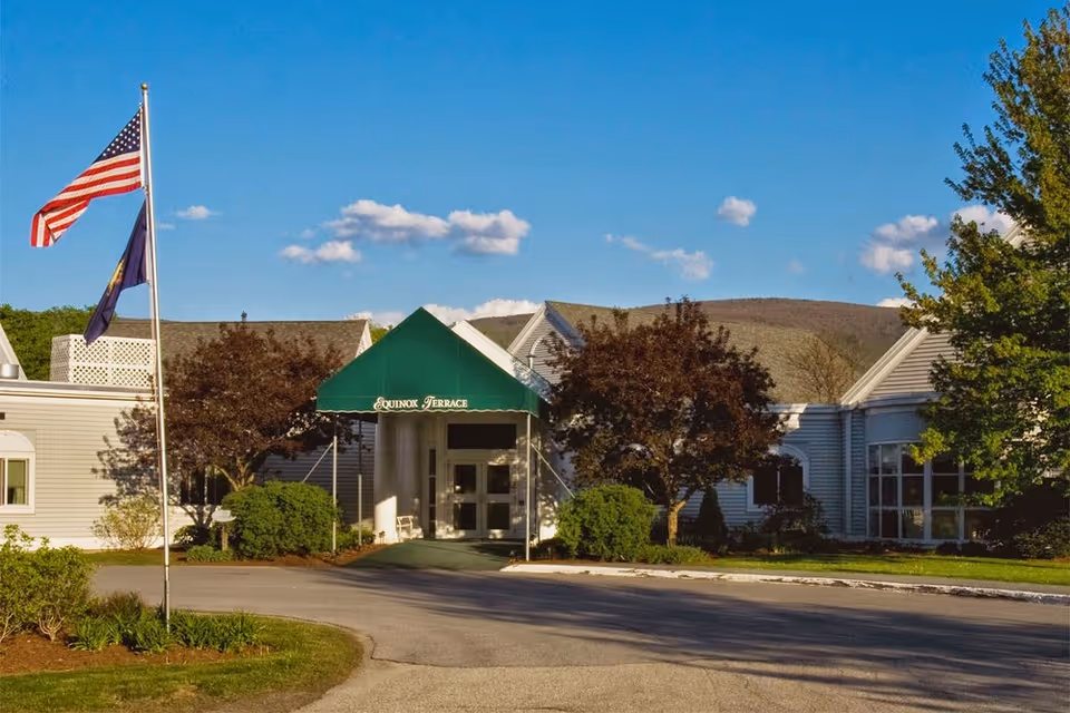 Exterior view of Equinox Terrace facility with a green entrance canopy displaying the facility name. The building is surrounded by trees and bushes, with a clear blue sky and a flagpole flying the American flag and another flag.