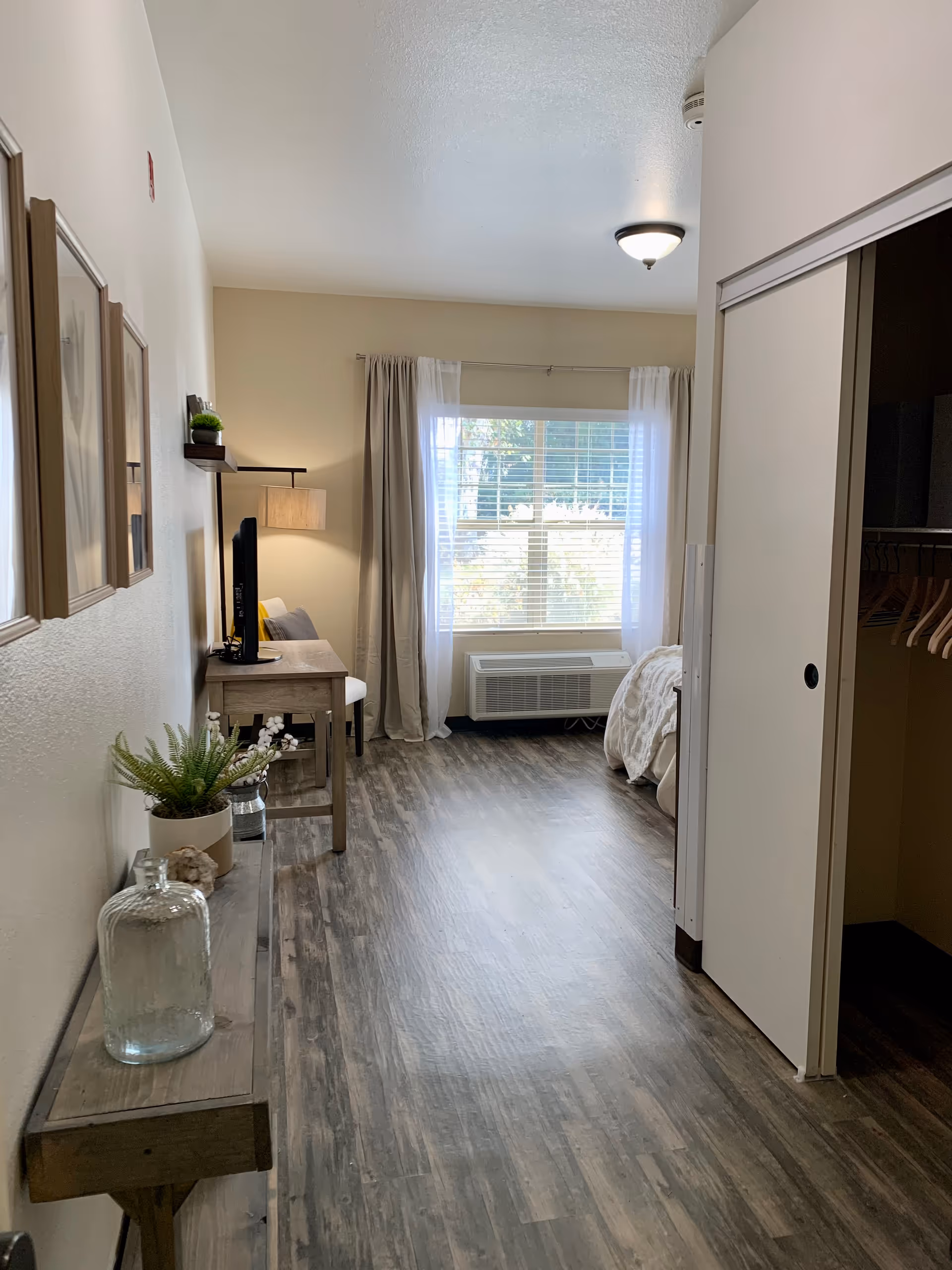 A bright and clean senior living bedroom with wood flooring, a window with curtains, a bed partially visible on the right, a closet with sliding doors, a small desk with a TV and chair, and a narrow wooden console table with decorative items along the left wall.