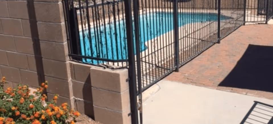 Gated outdoor pool area with a black metal fence, concrete block wall, pavers, and orange flowering plants.