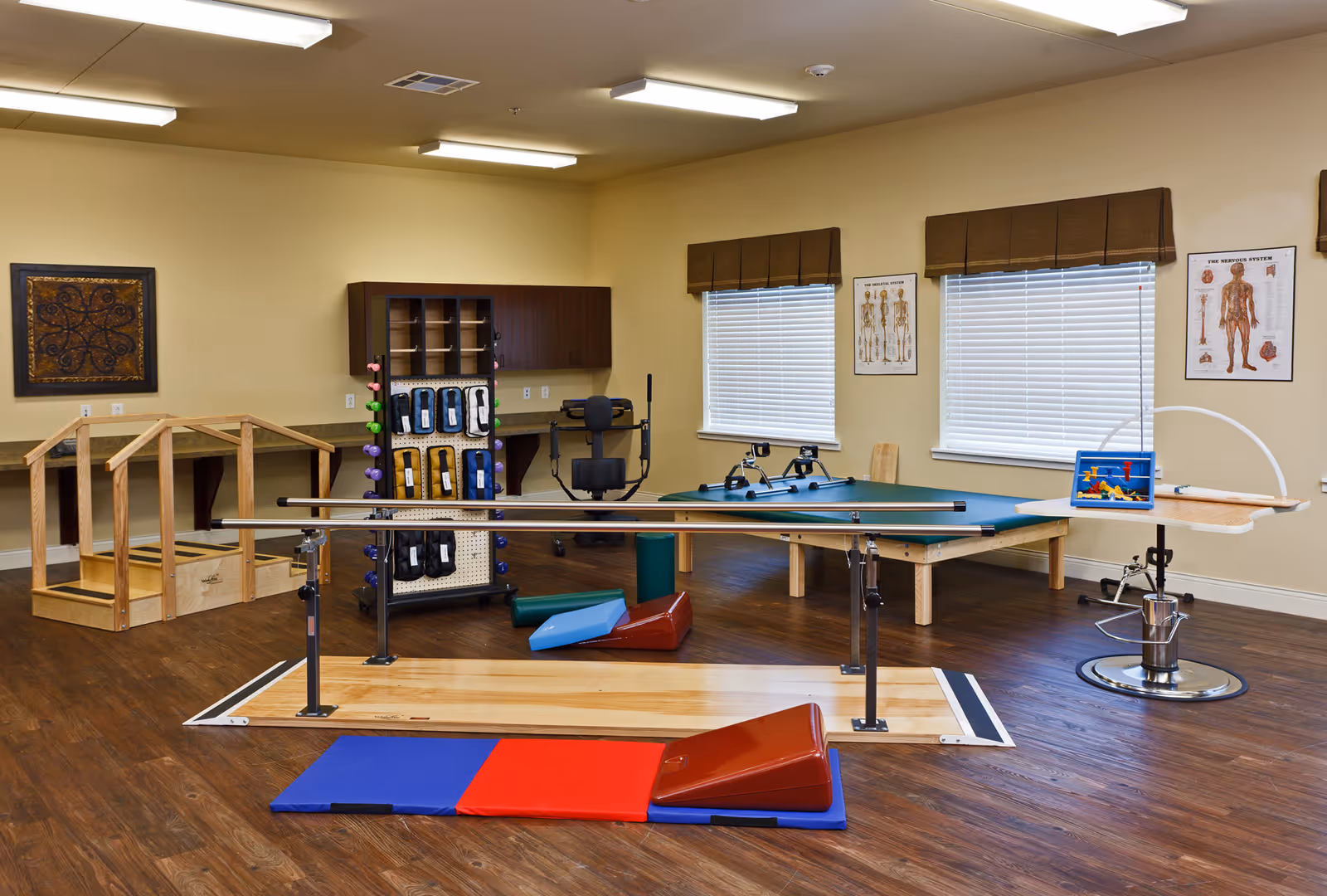 A rehabilitation therapy room with parallel bars for walking exercises, colorful foam wedges and mats on the wooden floor, a wooden step structure, exercise equipment, and anatomical charts on the walls. The room has two windows with blinds and brown valances, and a cabinet with various therapy tools.