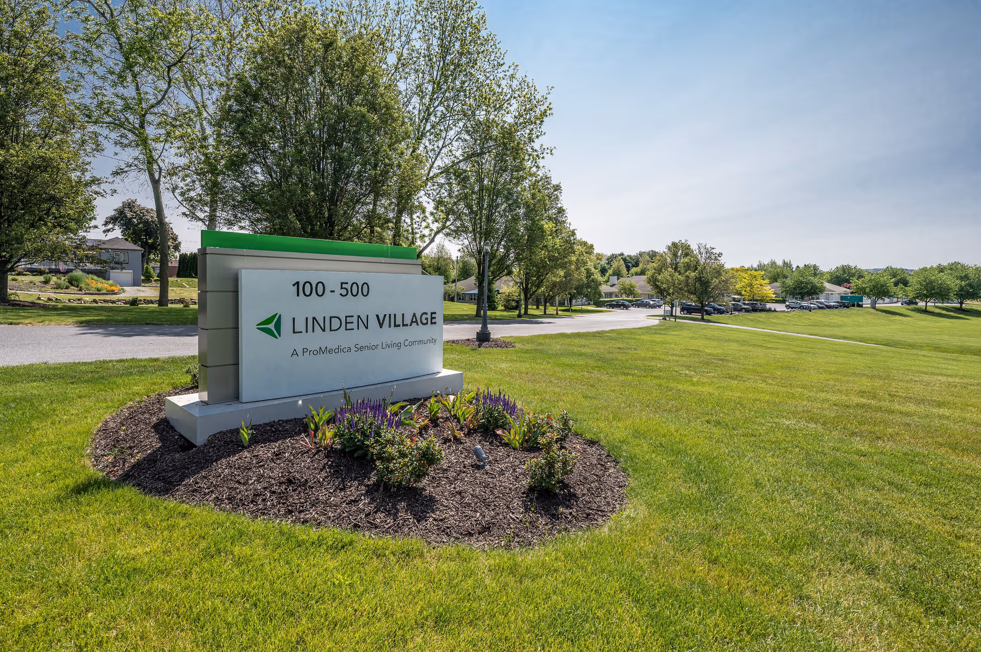 Outdoor view of the entrance sign for Linden Village, a ProMedica Senior Living Community, surrounded by green grass, trees, and a clear blue sky.