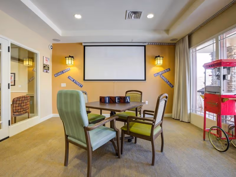 A cozy room with a table and four chairs arranged around it. On the table are blocks spelling out the word 'CINEMA'. The wall behind the table has a large white projector screen and two wall-mounted lantern-style lights. There are decorative street signs on the wall reading 'RODEO DRIVE', 'VINE STREET', 'HOLLYWOOD BLVD', and 'SUNSET BLVD'. To the right, there is a red popcorn machine near a large window with beige curtains. The room has beige carpet and light-colored walls.