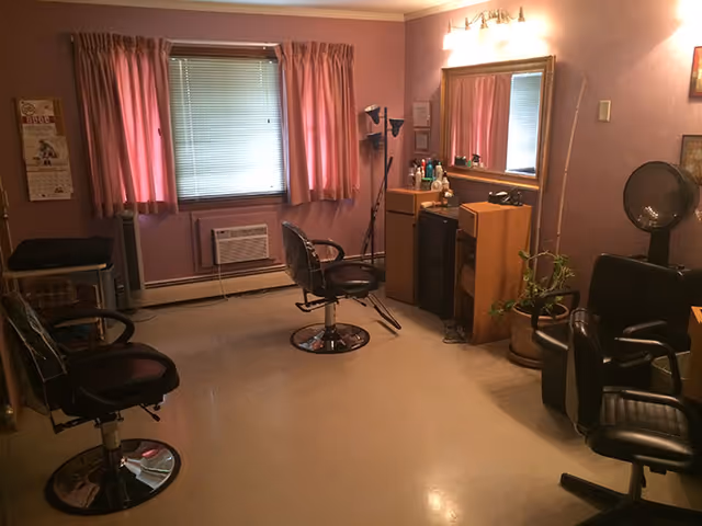 Interior of a small salon room with three black salon chairs, a large mirror on the wall, a window with pink curtains, an air conditioning unit below the window, and various salon equipment and plants.