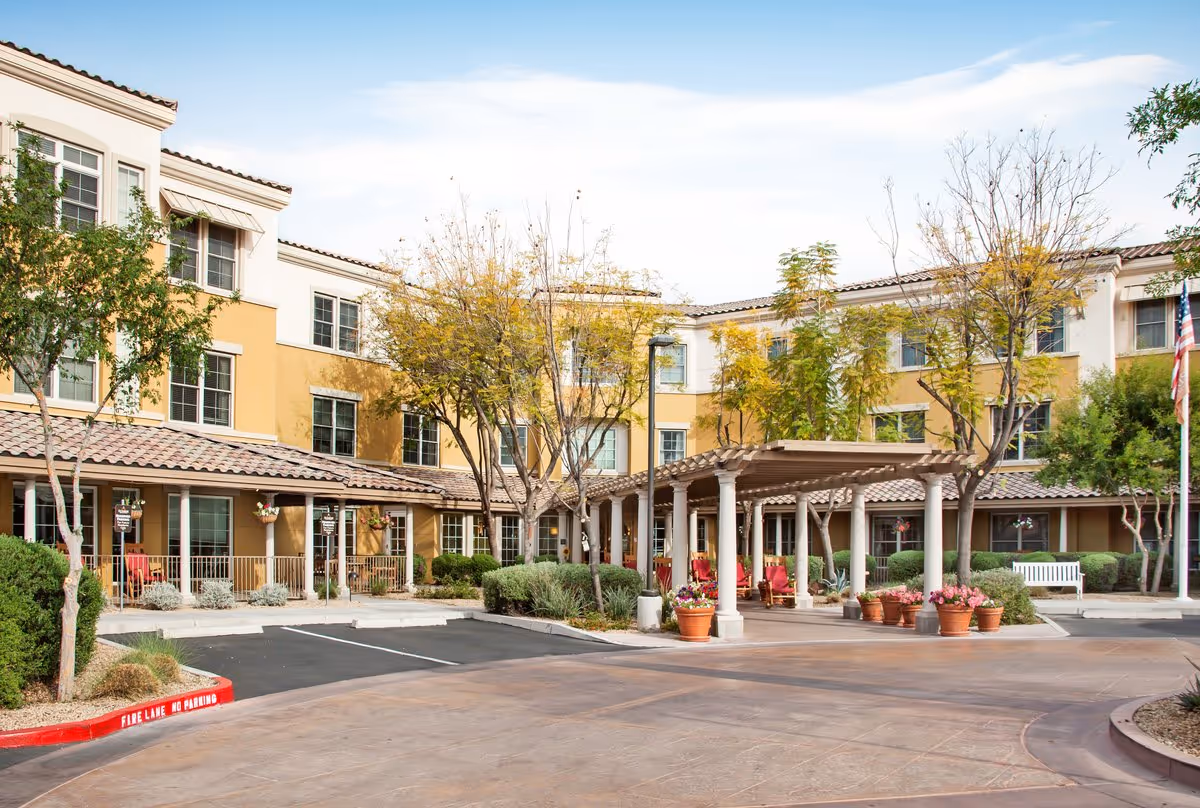 Front entrance of a three-story yellow stucco senior living building with a covered porte-cochere, potted plants, and trees.