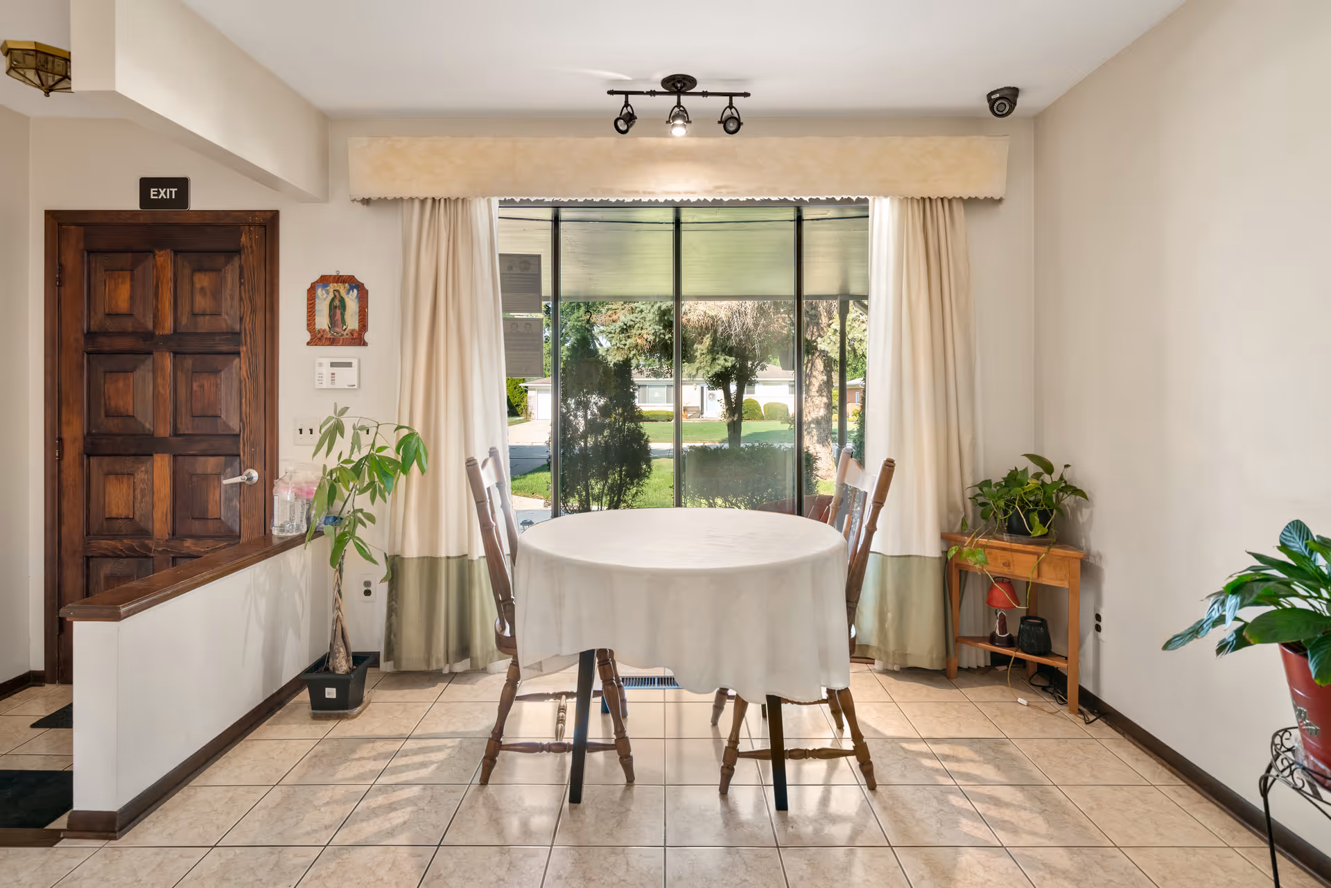 A small dining area with a round table covered with a white tablecloth and two wooden chairs. The room has large windows with cream and green curtains, letting in natural light and showing a view of trees and greenery outside. There are several potted plants around the room, a wooden door with an exit sign above it, and a small wooden side table with more plants and decorative items.