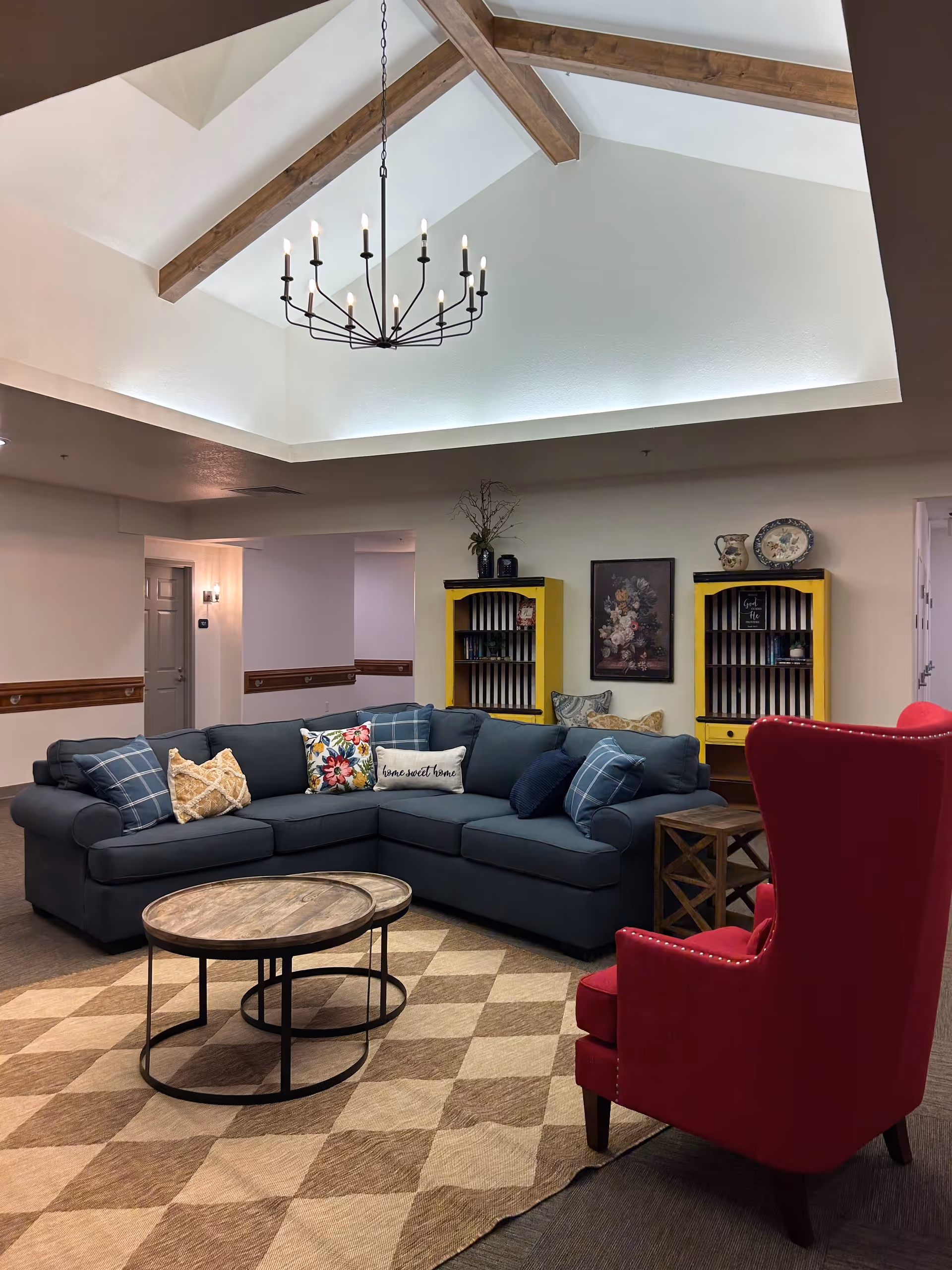 A cozy living room area with a large dark blue sectional sofa adorned with various patterned pillows, a round wooden coffee table, and a red upholstered armchair. The room features a high ceiling with exposed wooden beams and a modern chandelier. Two yellow bookshelves with decorative items and a floral painting are against the wall, and a checkered area rug covers the floor.