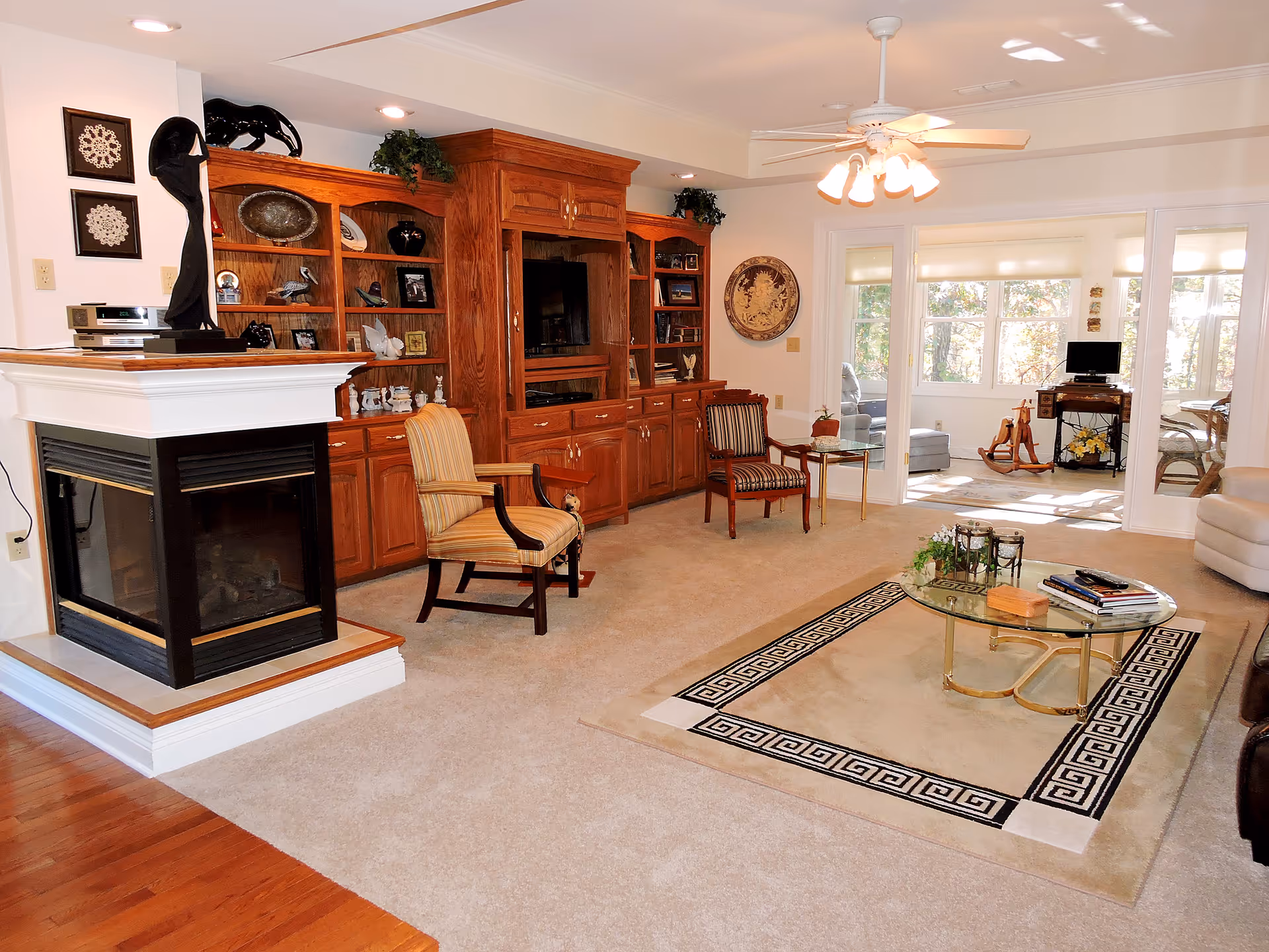 Spacious living room with a built-in wooden entertainment center, fireplace, chairs, and a glass coffee table on a decorative rug with a sunlit sitting area beyond.