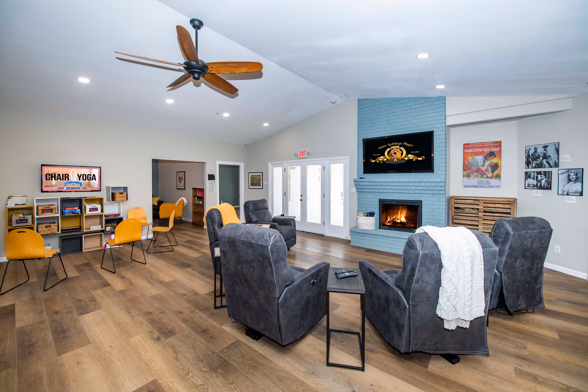 A spacious common living area with four gray armchairs arranged around a small black table in front of a blue brick fireplace with a fire burning. Above the fireplace is a mounted TV. To the left, there are yellow chairs and a shelving unit with baskets and a sign that reads 'CHAIR YOGA'. The room has wooden flooring, a ceiling fan with wooden blades, and several framed pictures on the walls.