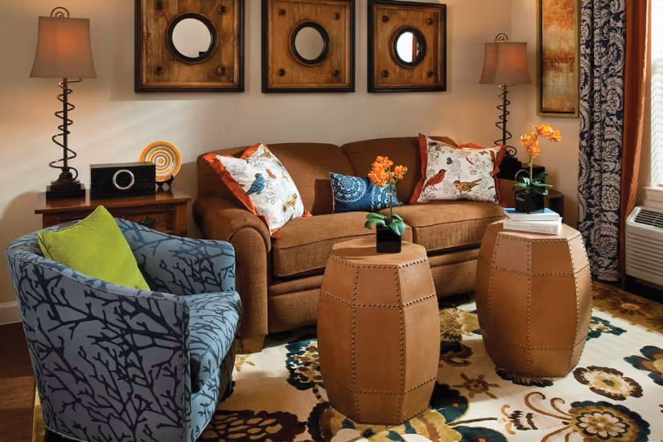 A cozy living room with a brown sofa adorned with bird-patterned pillows, a blue armchair with a green cushion, two brown leather ottomans serving as coffee tables with small orange flower arrangements on top, two side tables each with a lamp, decorative wall art including three square frames with circular mirrors, and a window with patterned curtains.