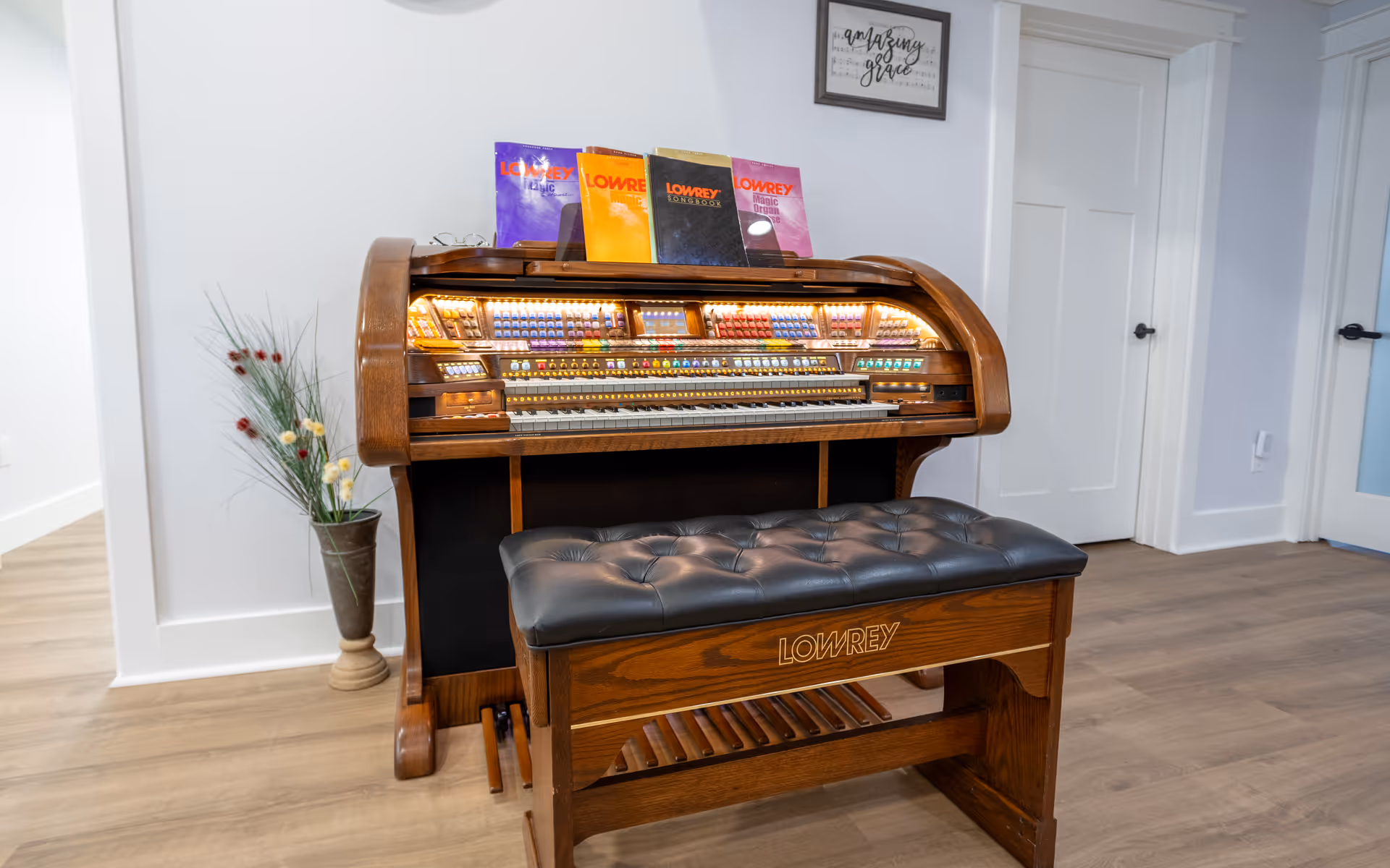 A vintage Lowrey organ with illuminated buttons and keys, accompanied by a cushioned bench with the Lowrey logo. The organ has several music books placed on top. It is situated in a room with light-colored walls, wooden flooring, a decorative vase with artificial flowers to the left, and two white doors in the background.