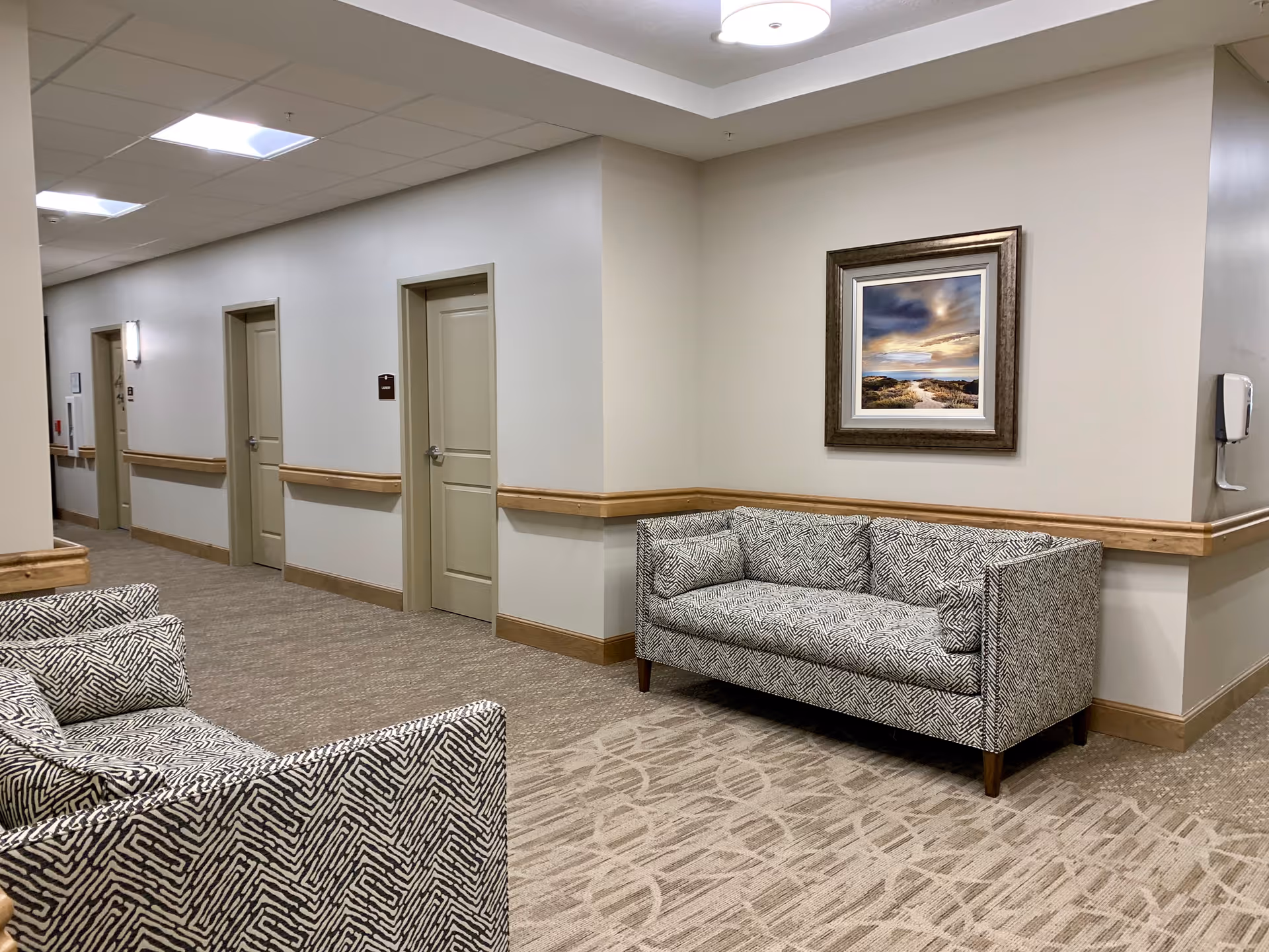 A hallway in an assisted living facility with beige walls and carpeted floors. There are three closed doors along the hallway, a patterned sofa against the wall on the right, and two matching patterned armchairs partially visible on the left. A framed landscape painting hangs above the sofa, and a hand sanitizer dispenser is mounted on the wall near the sofa.