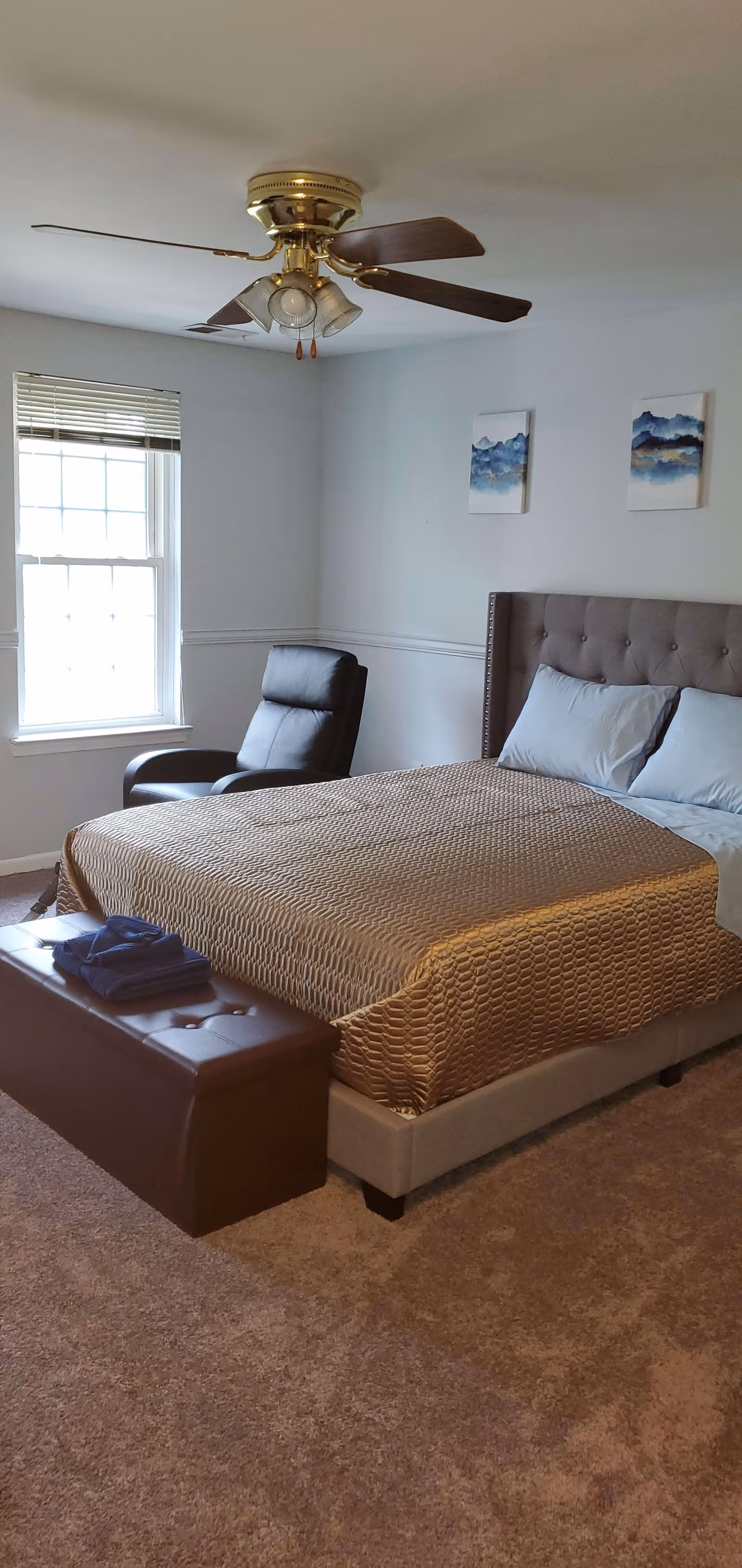 A bedroom with a large bed covered in a textured beige bedspread, two pillows, and a tufted headboard. At the foot of the bed is a brown storage bench with folded blue towels on top. To the left of the bed is a black recliner chair positioned near a window with white blinds. The walls are light-colored with two abstract blue and white paintings hanging above the bed. A ceiling fan with wooden blades and light fixtures is mounted on the ceiling.