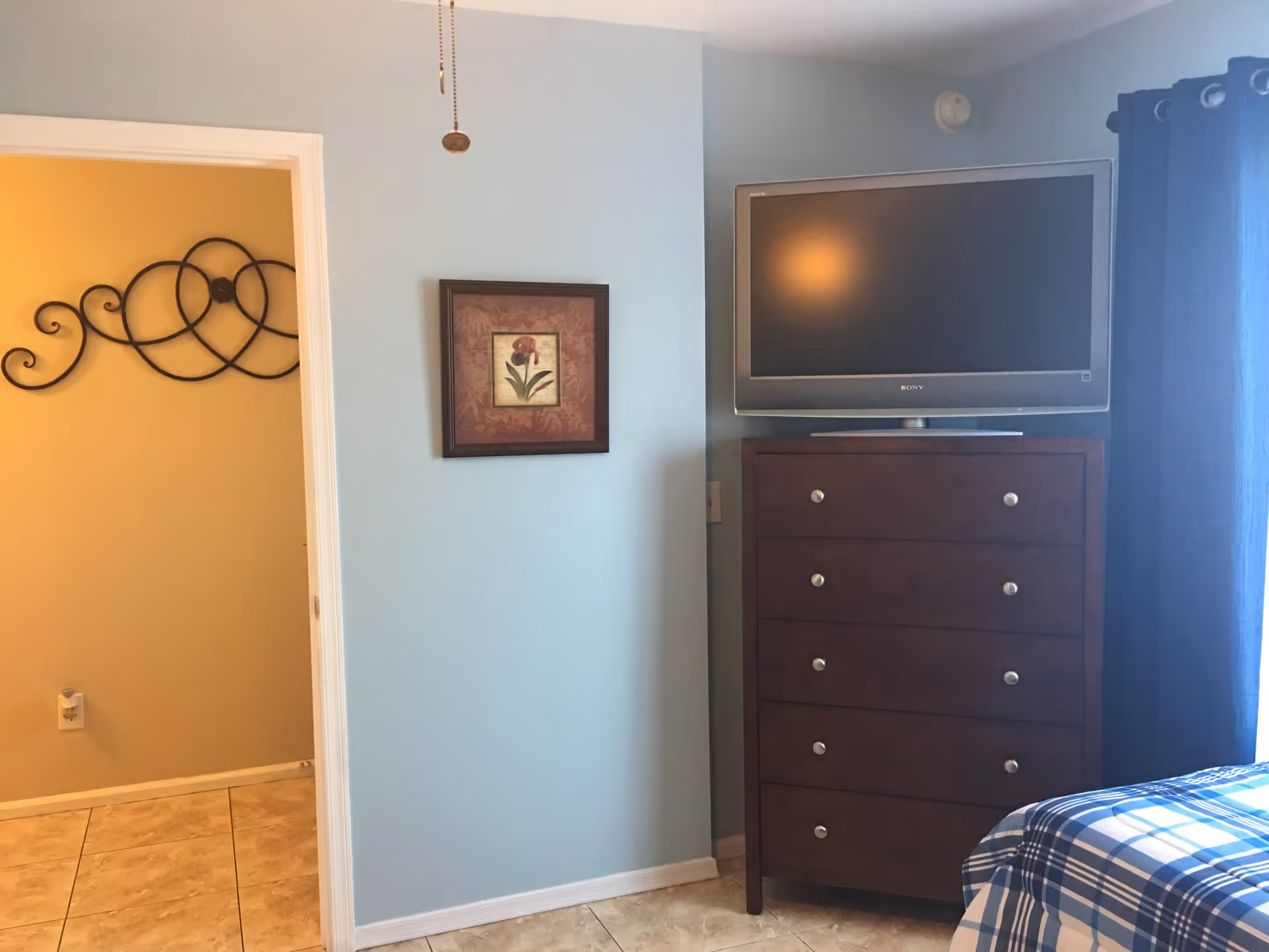 Bedroom interior showing a tall dark wood chest of drawers with a flat-screen TV, a light blue wall with framed art, and the corner of a plaid-covered bed.