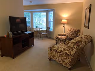 A cozy living room area with two patterned armchairs, a wooden TV stand with a flat-screen television, a floor lamp, and a small round table with two chairs near a bay window letting in natural light.