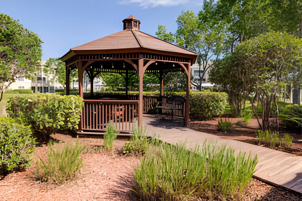 A wooden gazebo with a peaked roof situated in a landscaped garden area with green shrubs, trees, and a paved walkway leading to it under a clear blue sky.