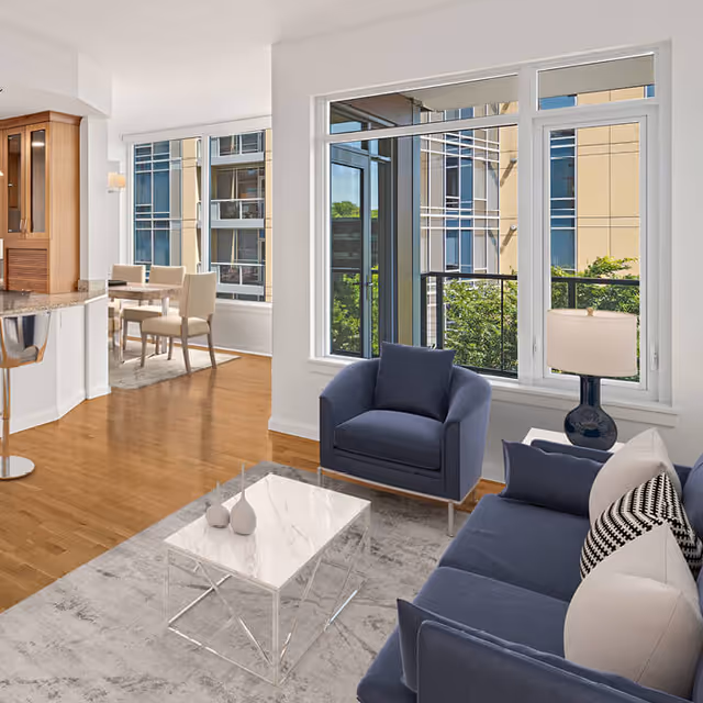 Open-plan living area with a navy sofa and armchair, marble-top coffee table, large windows, and a dining area in the background.