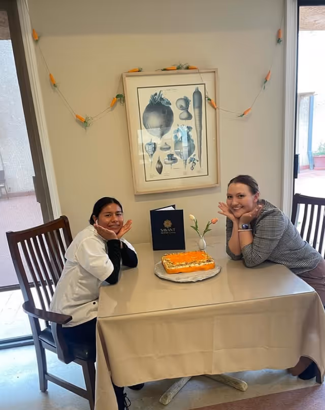 Two women sitting at a table covered with a beige tablecloth, smiling and resting their faces on their hands. On the table is a rectangular cake with orange and white frosting, a small vase with two tulips, and a Savant of Santa Monica brochure. Behind them is a wall with a framed botanical print and a string of carrot-shaped decorations. A sliding glass door is visible to the right.