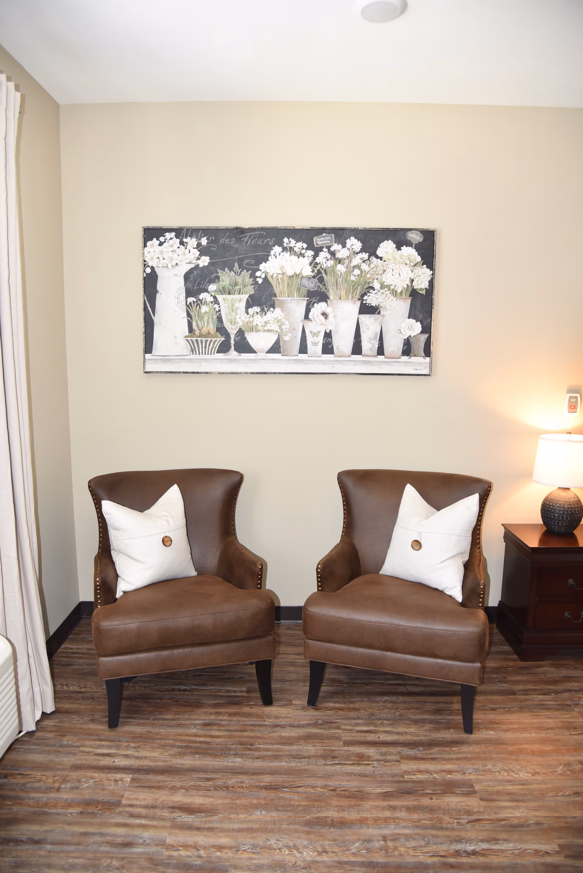 Two brown leather armchairs with white pillows are placed side by side on a wooden floor. Behind the chairs, a wall features a large framed artwork depicting white flowers in various vases. To the right of the chairs, there is a dark wooden side table with a textured lamp emitting warm light. Light-colored curtains are partially visible on the left side of the image.