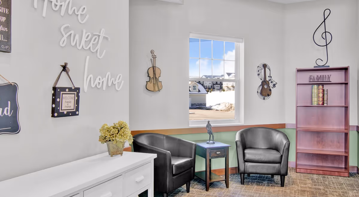 Cozy seating area with two black armchairs, a small table and lamp, decorative wall art saying 'Home sweet home', a bookshelf labeled 'FAMILY', and a window showing houses outside.