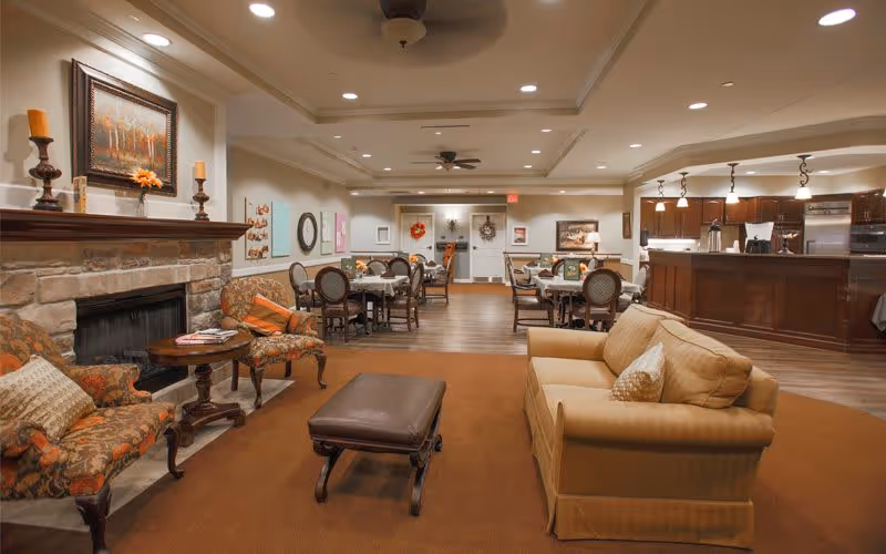 A spacious and warmly lit common area in a senior living facility featuring a stone fireplace with candles and a painting above it, comfortable upholstered chairs and a sofa with cushions, a brown ottoman, several dining tables with chairs, and a kitchen area with dark wood cabinetry and stainless steel appliances in the background.