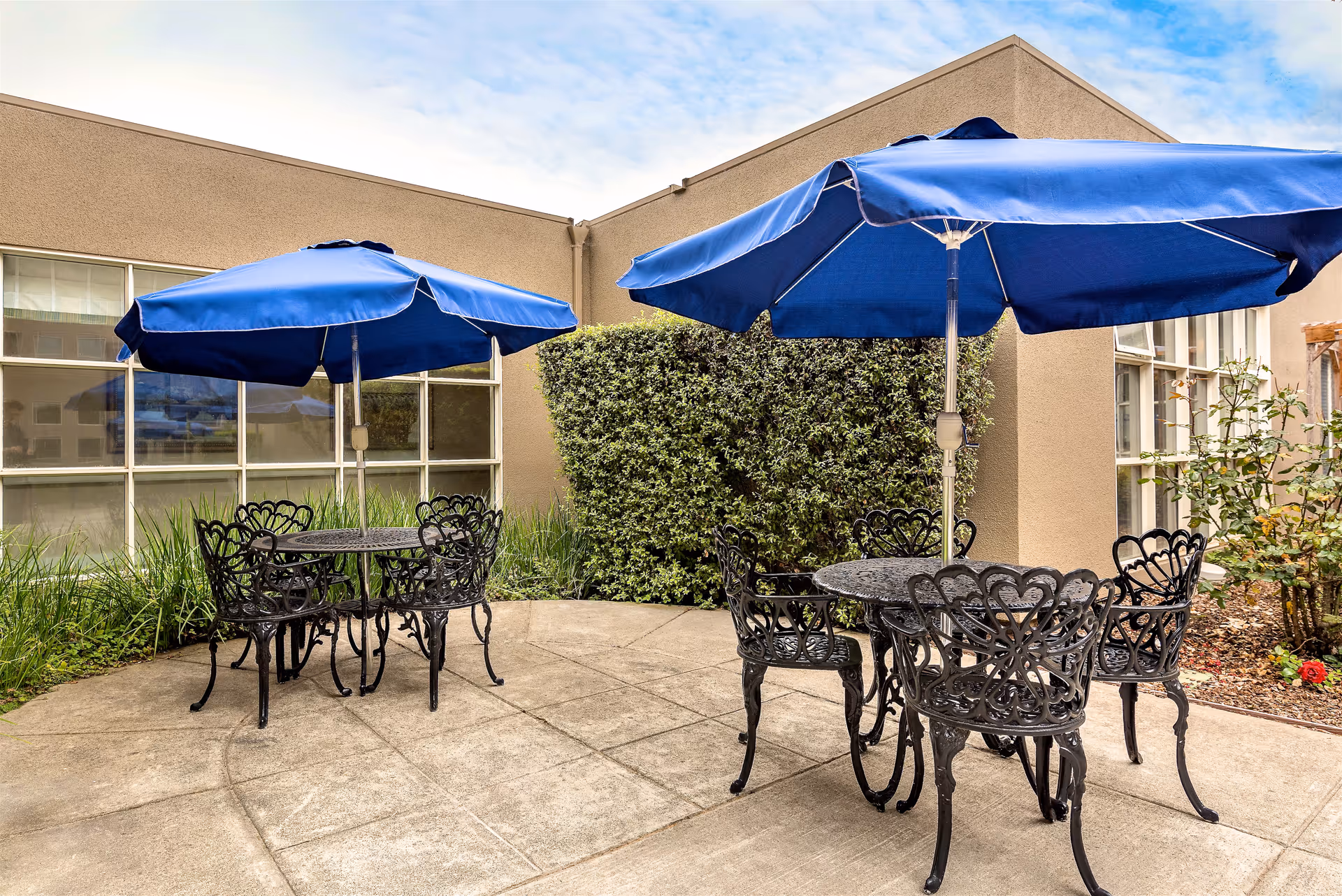 Outdoor patio area at Mission Villa Senior Living featuring two black metal tables with intricate designs, each surrounded by matching chairs. Each table has a large blue umbrella providing shade. The patio is paved with concrete and bordered by bushes and plants. The building's beige exterior walls and windows are visible in the background under a partly cloudy sky.