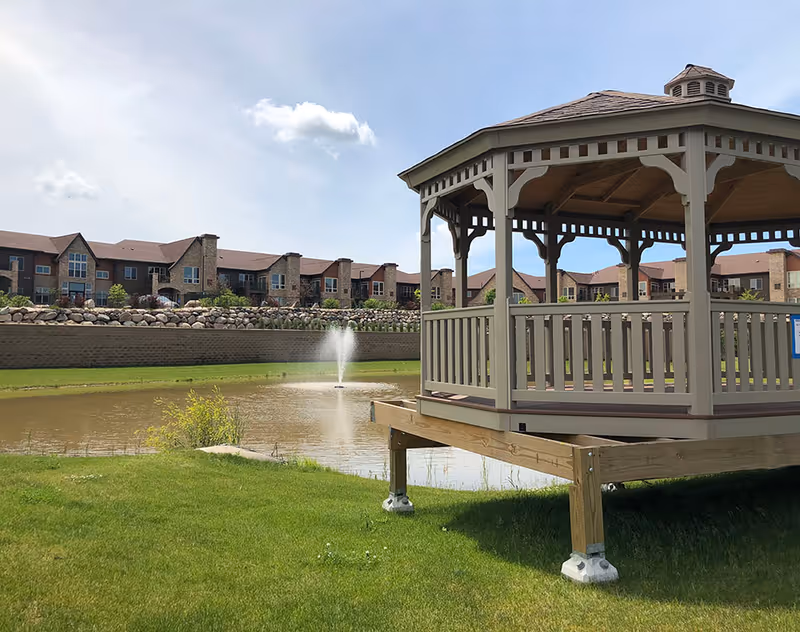 Outdoor view of a senior living facility featuring a wooden gazebo on a grassy area next to a pond with a water fountain. In the background, there are multi-story residential buildings under a partly cloudy sky.