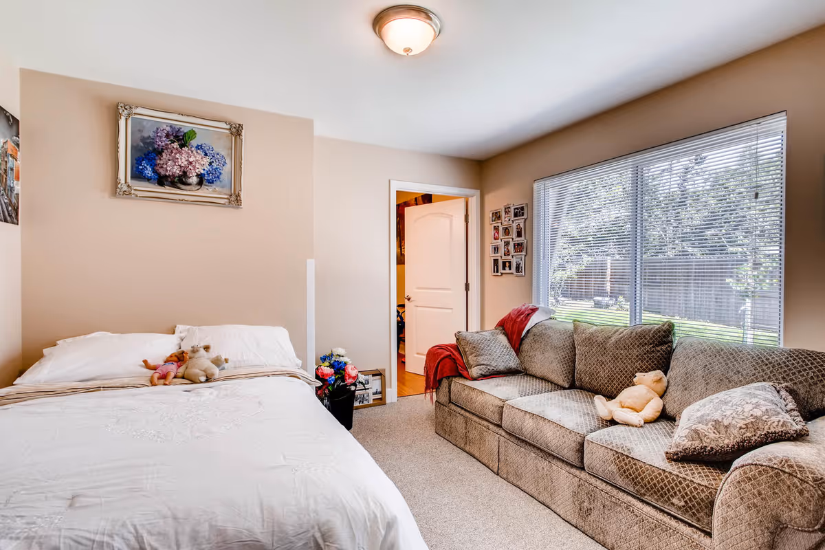A cozy bedroom with a white bed on the left side, decorated with two stuffed toys. On the right side, there is a large patterned sofa with cushions and a teddy bear. A window with blinds lets in natural light, and a door is open to another room. The walls are beige, with a framed floral painting above the bed and a collage of photos on the wall near the window.
