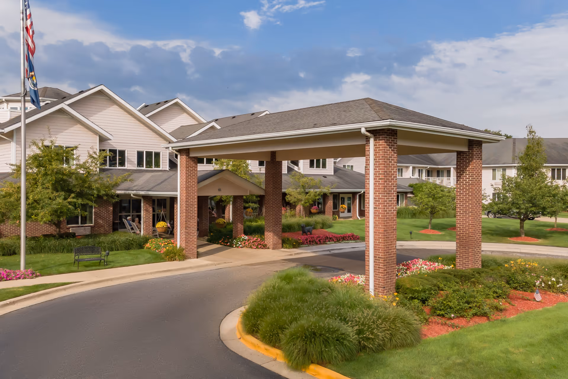 Entrance of StoryPoint Grand Rapids West senior living facility with a covered driveway supported by brick columns, surrounded by well-maintained landscaping including green grass, bushes, flowers, and trees under a partly cloudy sky.
