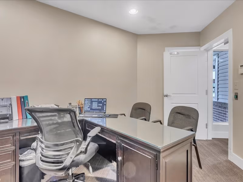 An office space with a large dark wooden desk topped with glass, a black ergonomic office chair, two guest chairs, a laptop, and office supplies. The room has beige walls, a white door that is open showing an exterior view, and carpeted flooring.