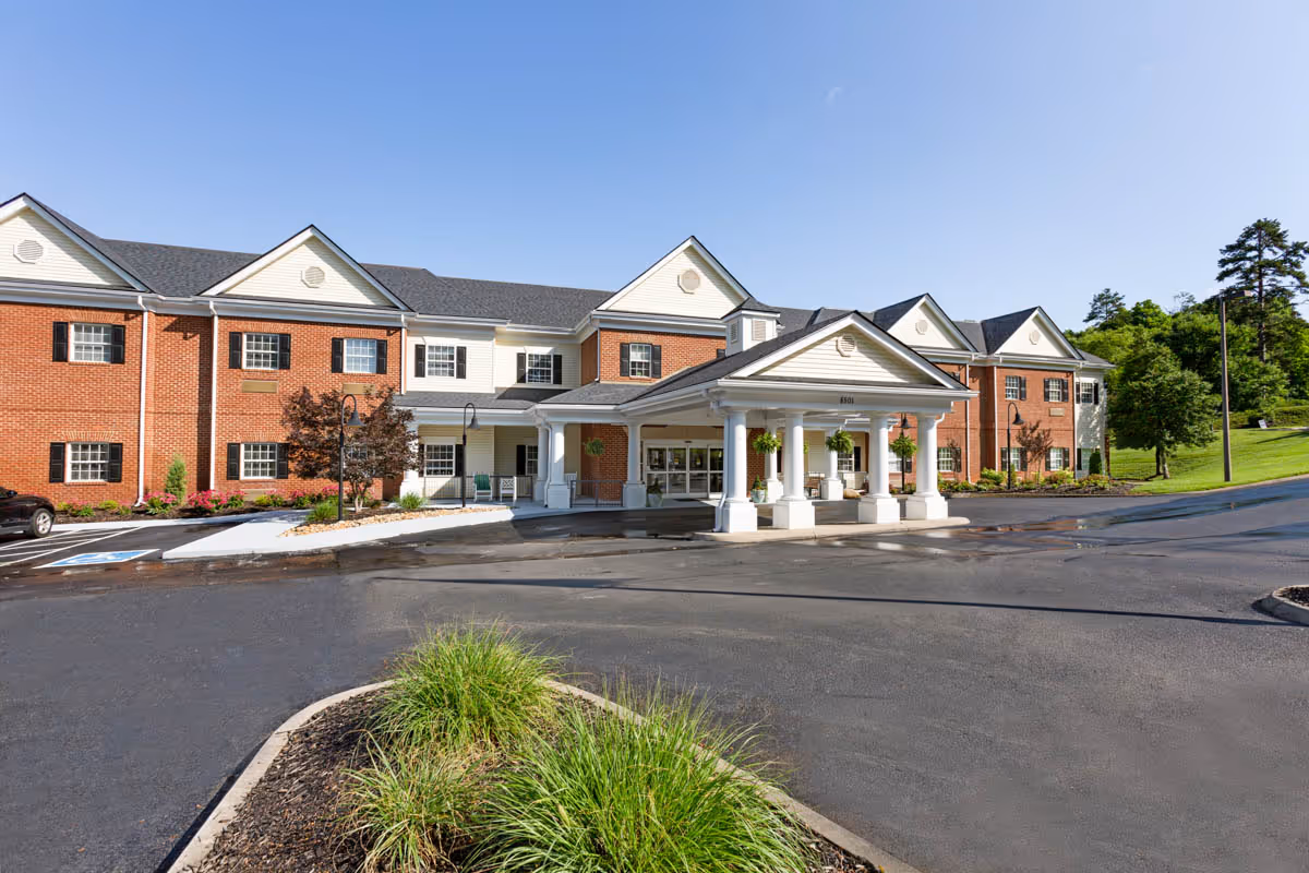 Front entrance of a two-story red brick senior living facility with a covered porte-cochère and landscaped driveway.