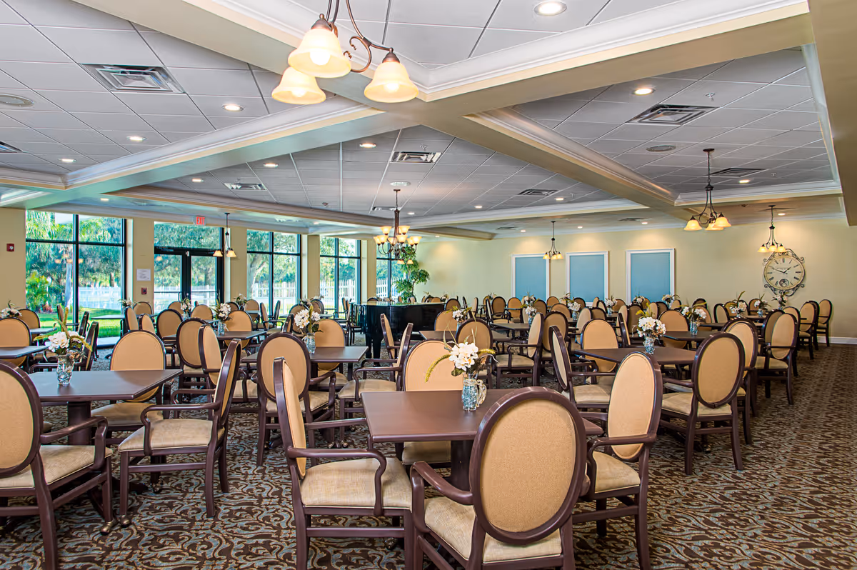 Spacious dining room with multiple tables and cushioned chairs arranged neatly. Each table has a small floral centerpiece. Large windows on one side allow natural light to brighten the room, and decorative ceiling lights hang overhead. The room has a patterned carpet and light-colored walls with a large clock on one wall.