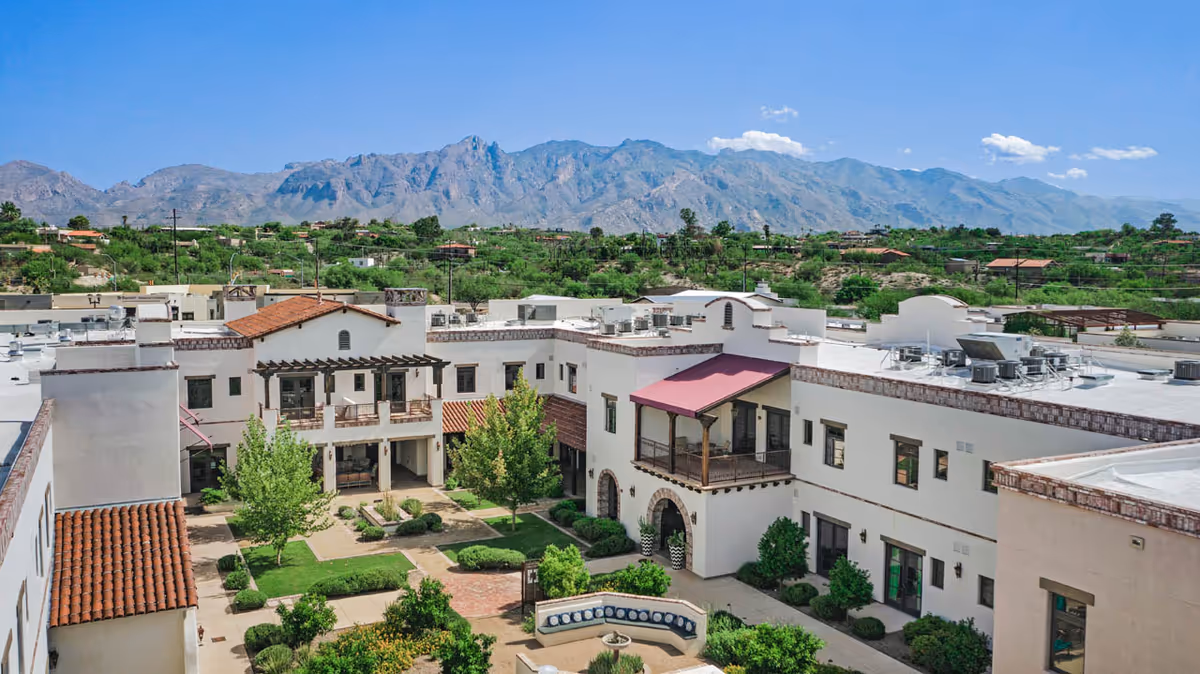 View of a senior living facility courtyard with green trees, shrubs, and walkways surrounded by a two-story white building with red tile roofs and balconies, set against a backdrop of mountains under a clear blue sky.