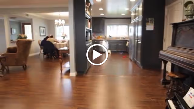 Interior view of a senior living facility showing a spacious area with wooden flooring. On the right side, there is a piano with a stool. In the background, there is a kitchen area with cabinets and a window. To the left, there is a dining area with people seated around a table under a chandelier.