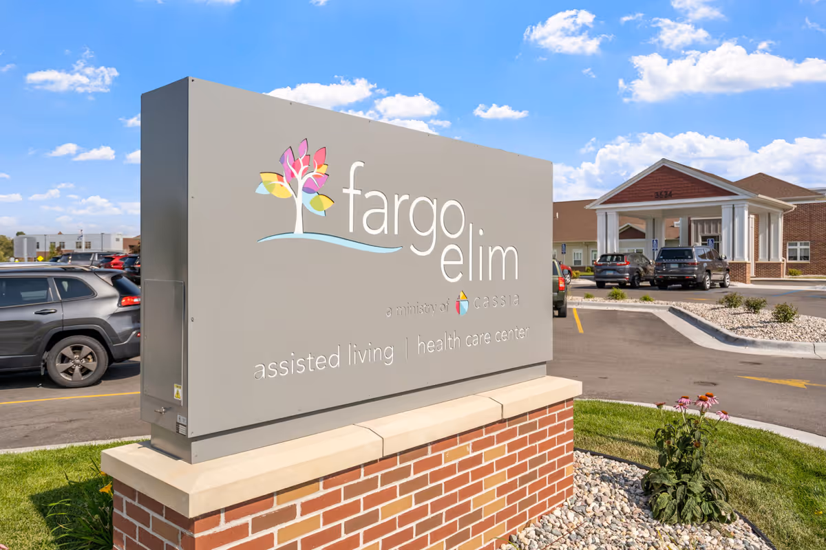Outdoor view of the Fargo Elim Health Care Center sign with a colorful tree logo, located on a brick base. In the background, there is a parking lot with several cars and the entrance to the health care center building under a blue sky with some clouds.
