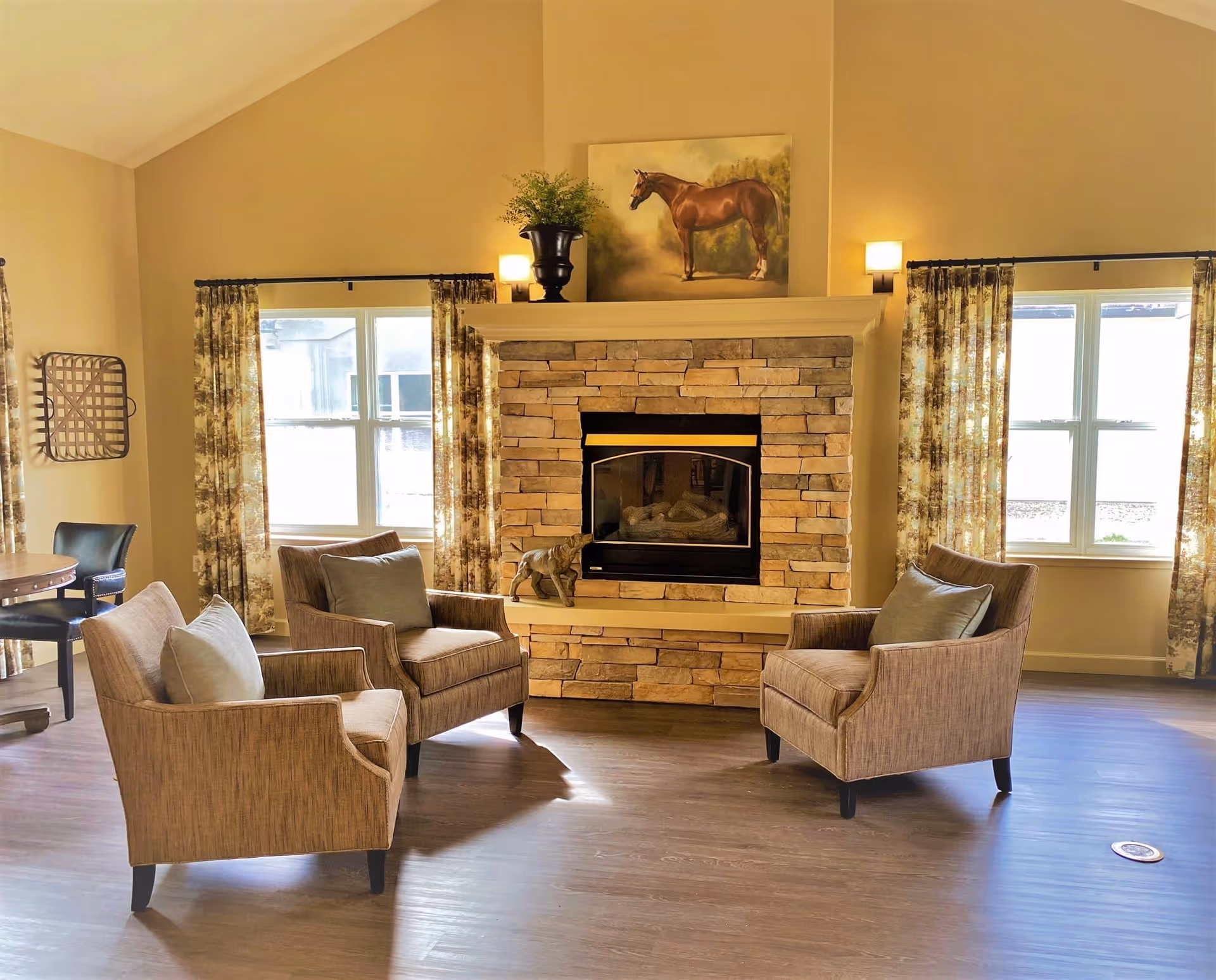 A cozy living room with three beige armchairs arranged around a stone fireplace. Above the fireplace is a painting of a horse and a potted plant. Two windows with patterned curtains flank the fireplace, allowing natural light into the room. The floor is wooden, and the walls are painted a warm beige color.