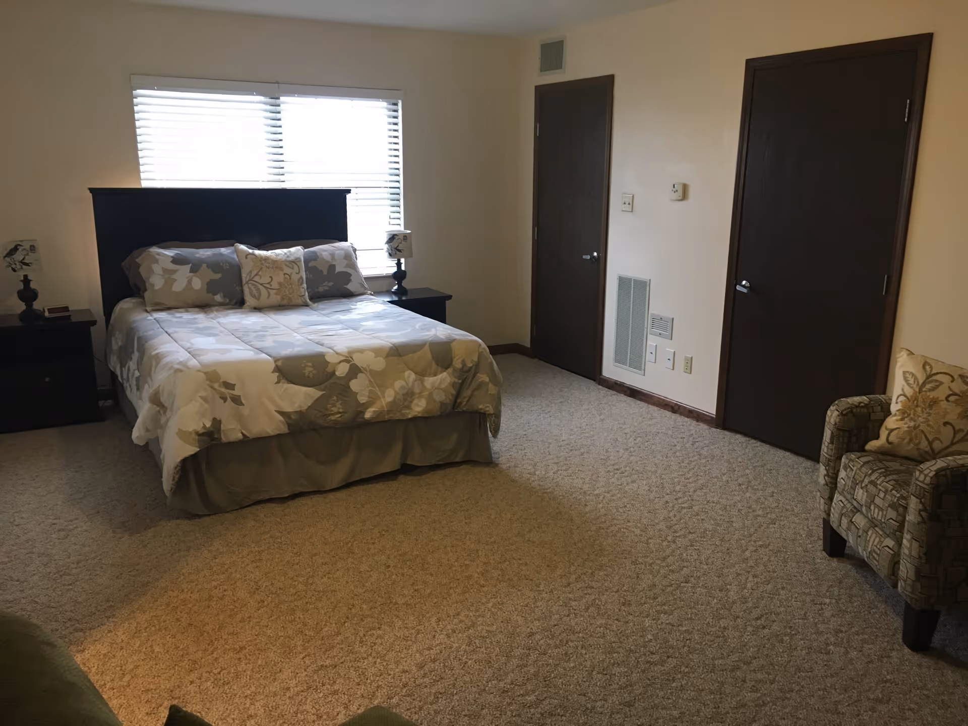 A bedroom with a large bed covered in a floral patterned comforter and pillows. There are two dark wooden nightstands with matching lamps on either side of the bed. A window with blinds is behind the bed. The room has beige carpet, two closed dark wooden doors on the right wall, and a patterned armchair with a decorative pillow in the corner.