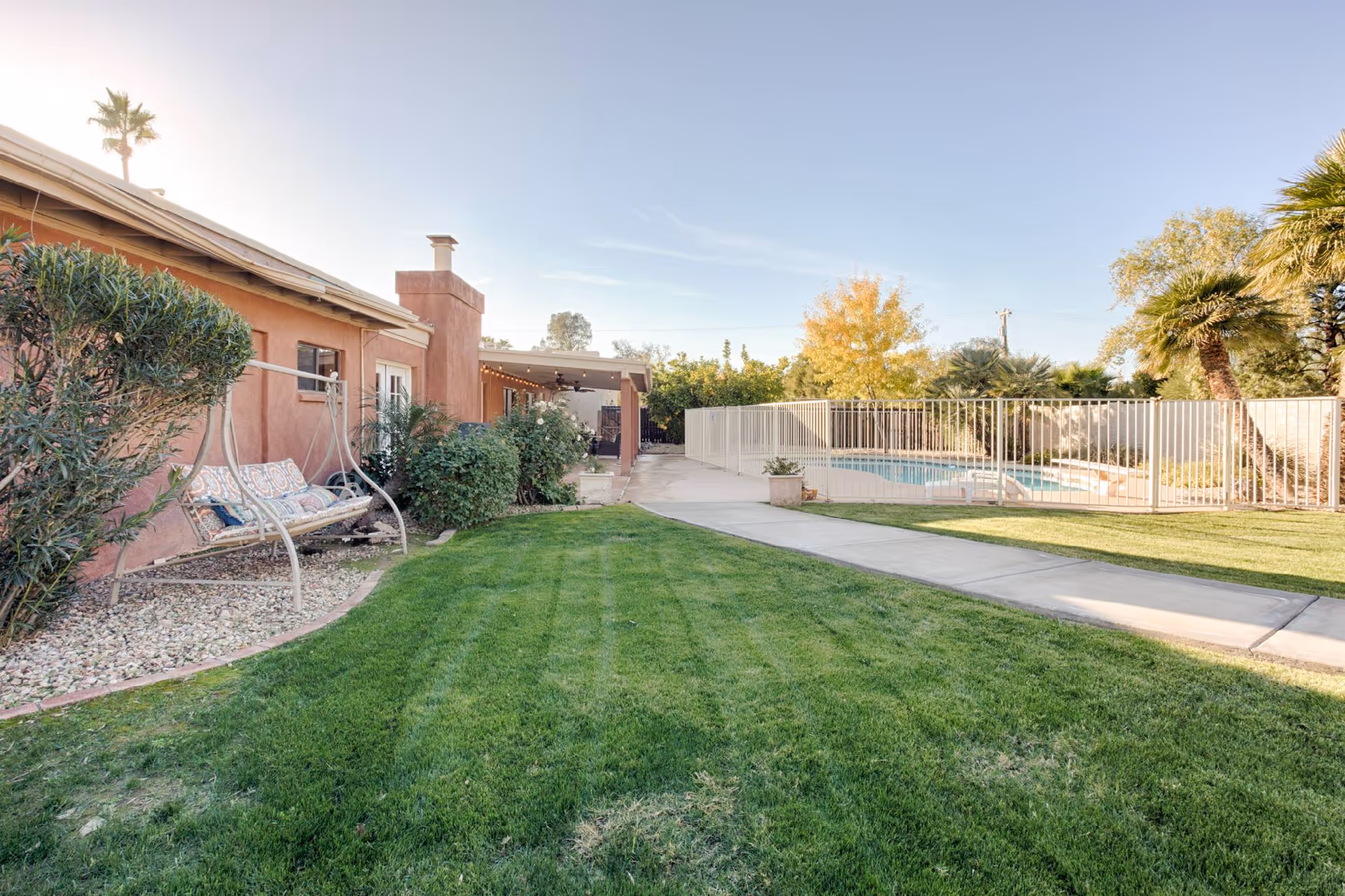 Outdoor view of a senior living facility's backyard area with a green lawn, a swing bench with cushions, a paved walkway, and a fenced swimming pool surrounded by trees and plants under a clear sky.
