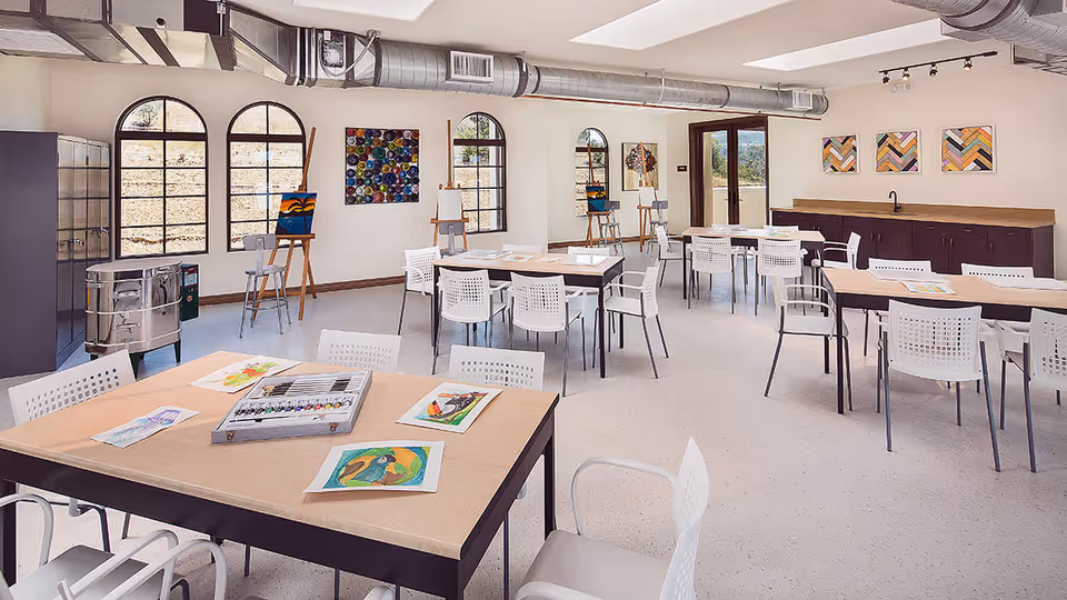 A bright and spacious art room with multiple tables and white chairs arranged for group activities. The tables have watercolor paintings and art supplies on them. The room features large arched windows, colorful artwork on the walls, and easels with paintings. There is a counter with cabinets along one wall and a door leading outside.